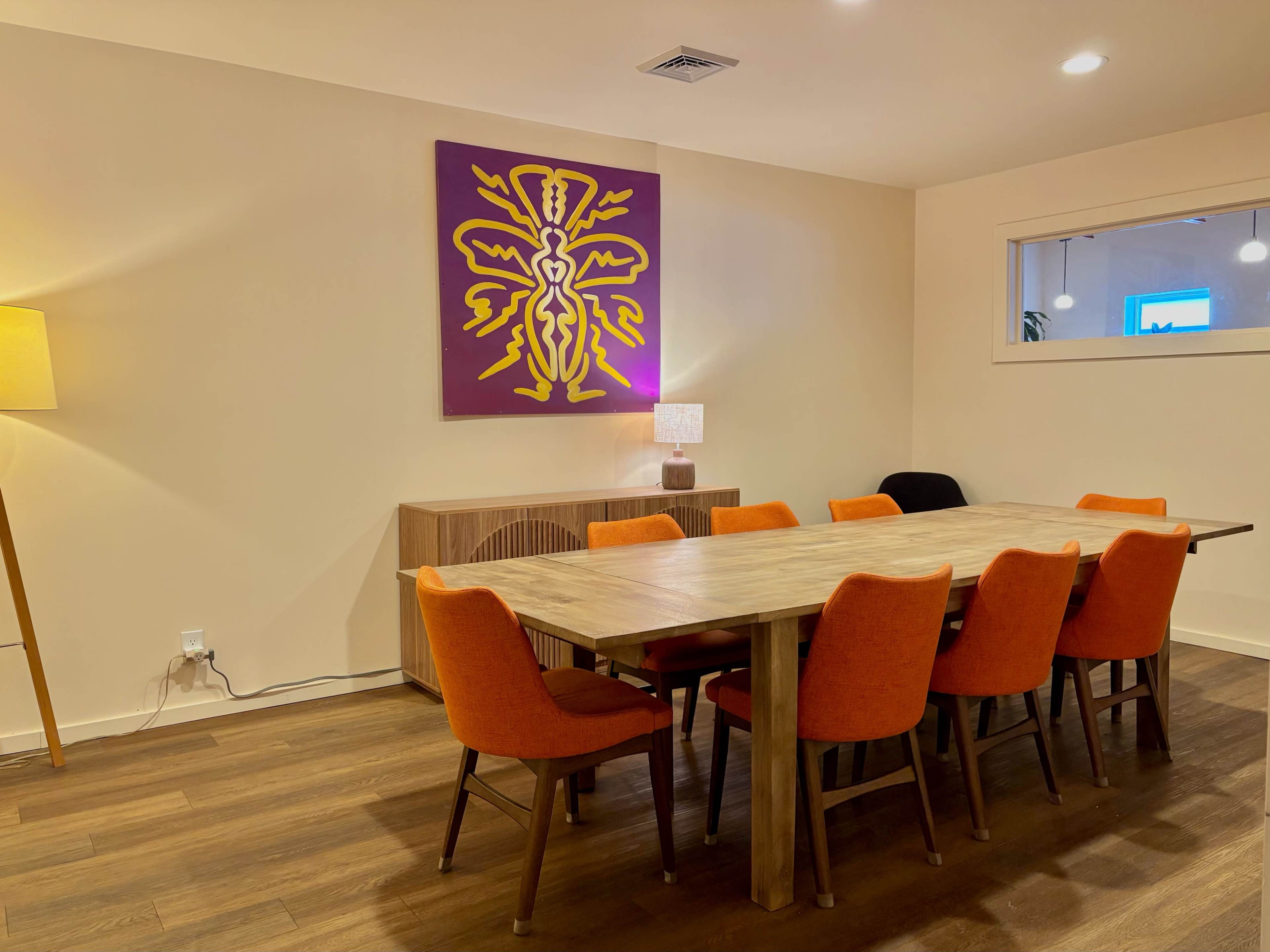 The image depicts a dining room with a large wooden table surrounded by orange chairs, a sideboard, and a brightly colored artwork hanging on the wall.
