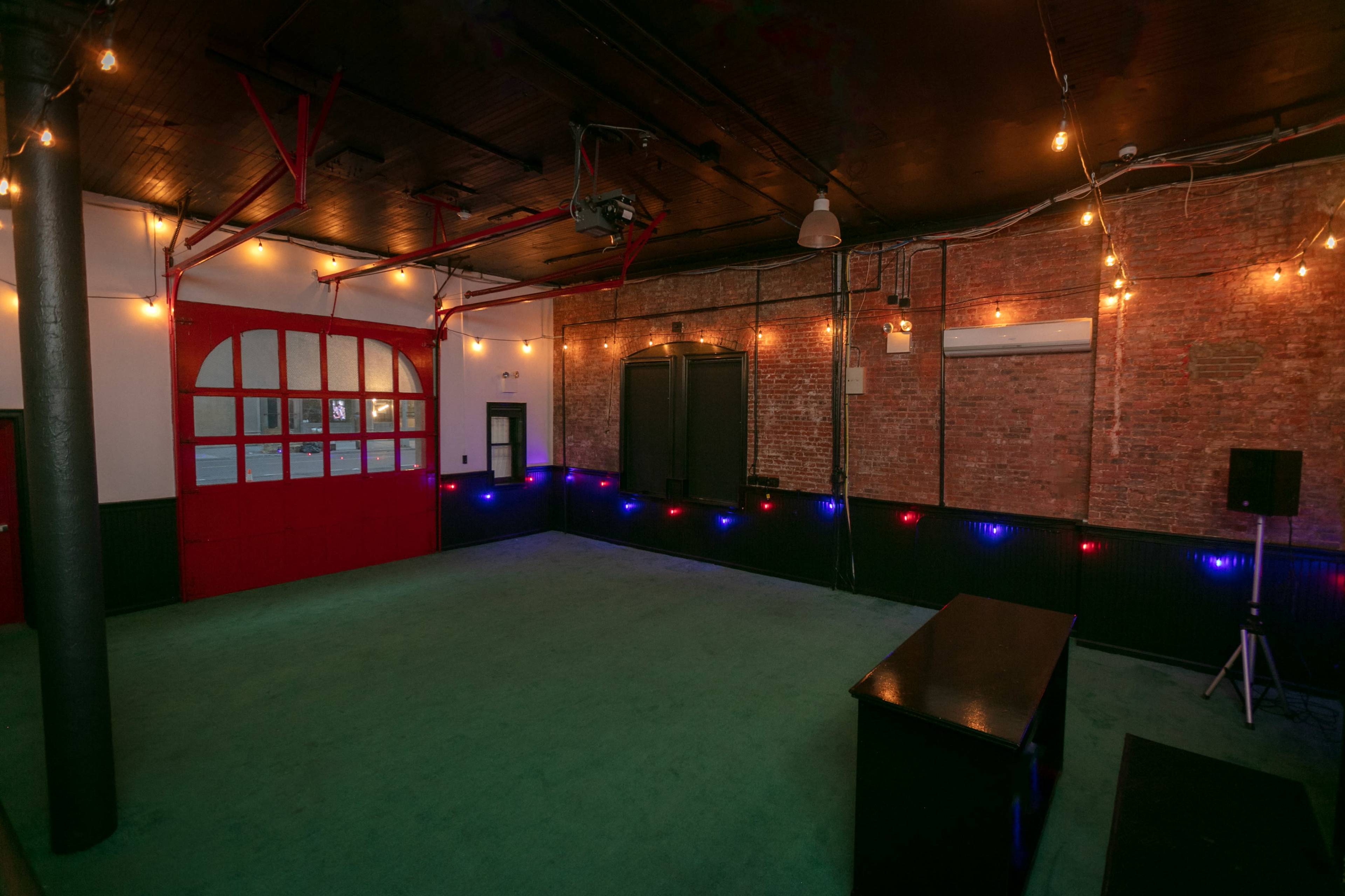 The image shows an empty, dimly lit room with exposed brick walls, a small stage, and string lights along the perimeter.