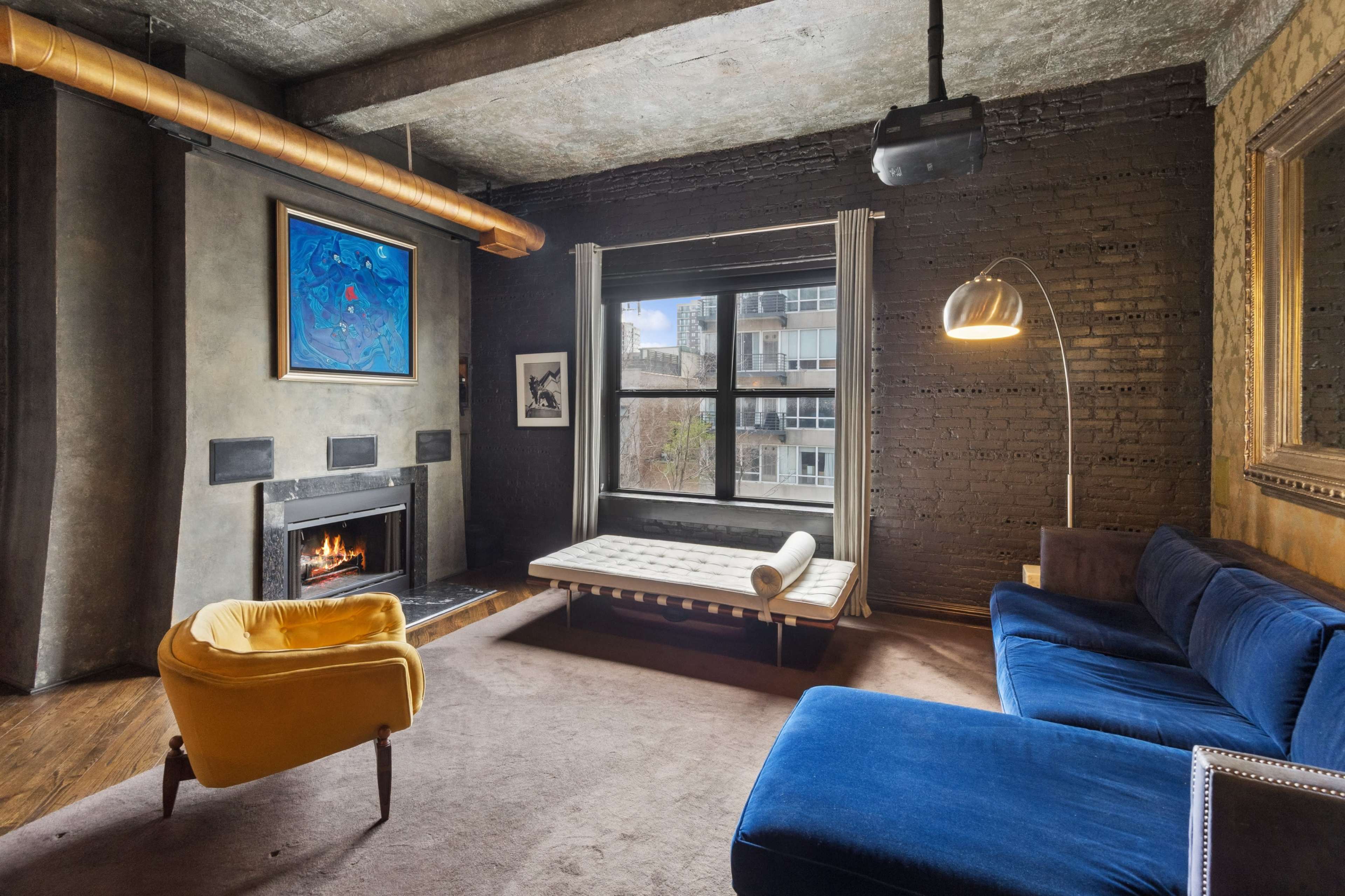 The living room features a blue sofa, a yellow chair, a fireplace, and a large window overlooking an urban landscape.