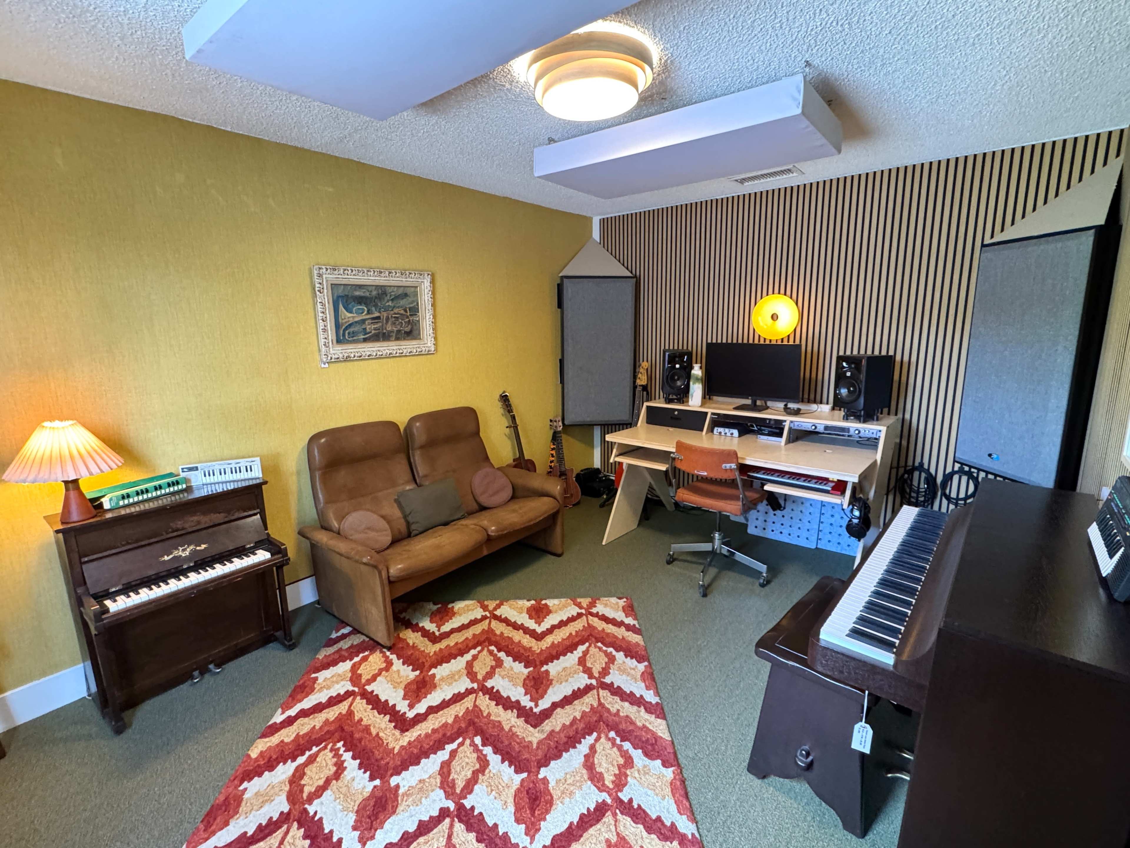 The room features a cozy music studio with a brown couch, a piano, and a keyboard, along with a desk setup equipped with audio monitors and a computer.