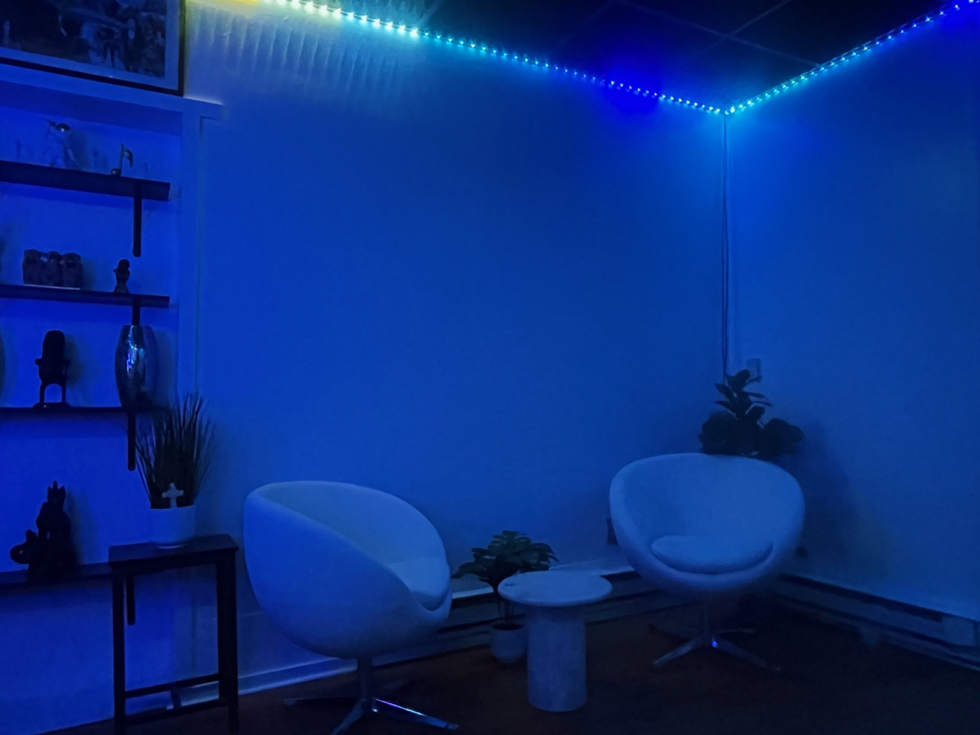 The image shows a dimly lit room featuring two white chairs and a small round table, illuminated by blue LED strip lights along the walls.