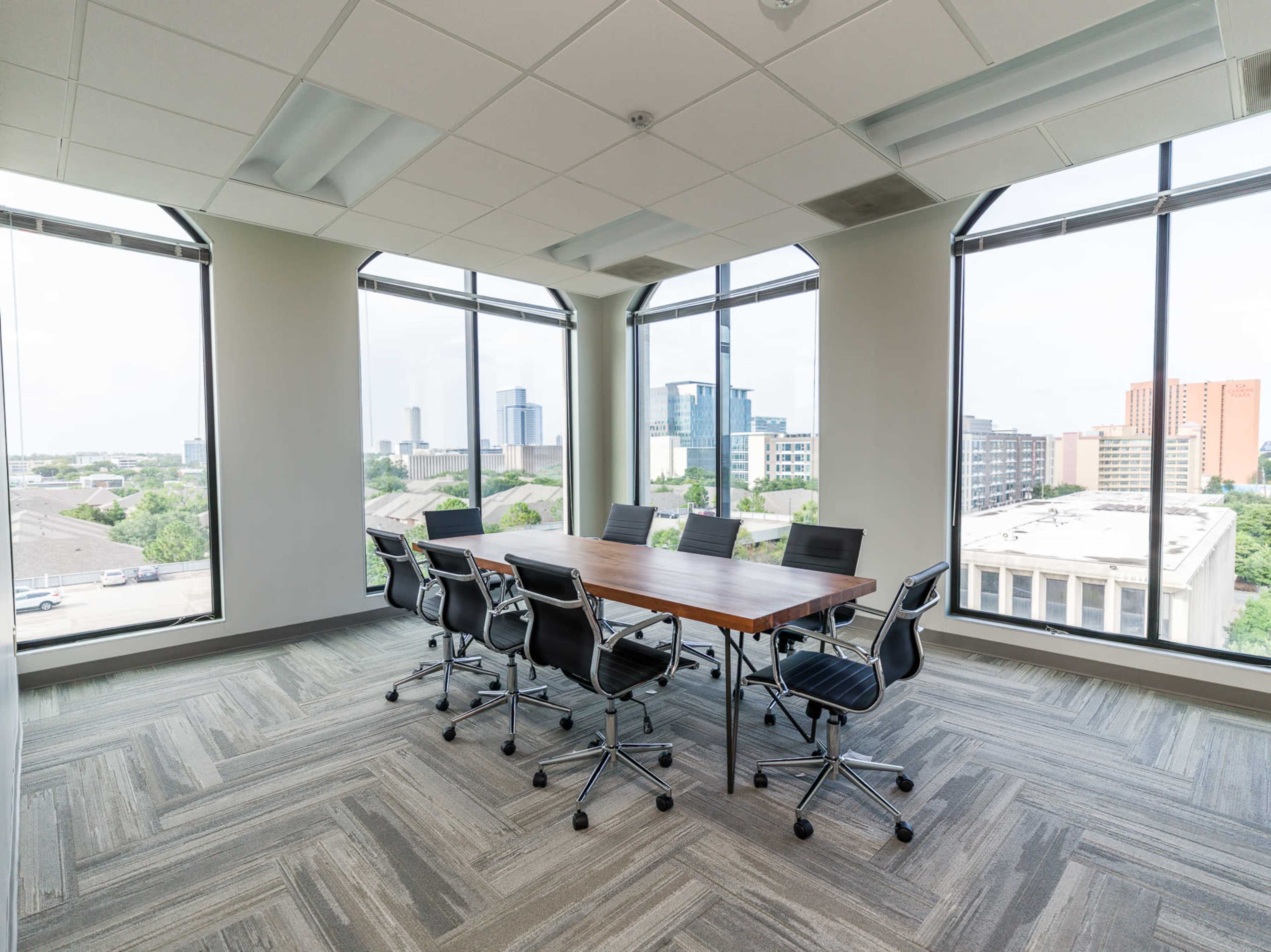 A modern conference room features a large table surrounded by black office chairs, with large windows offering a view of the city skyline.