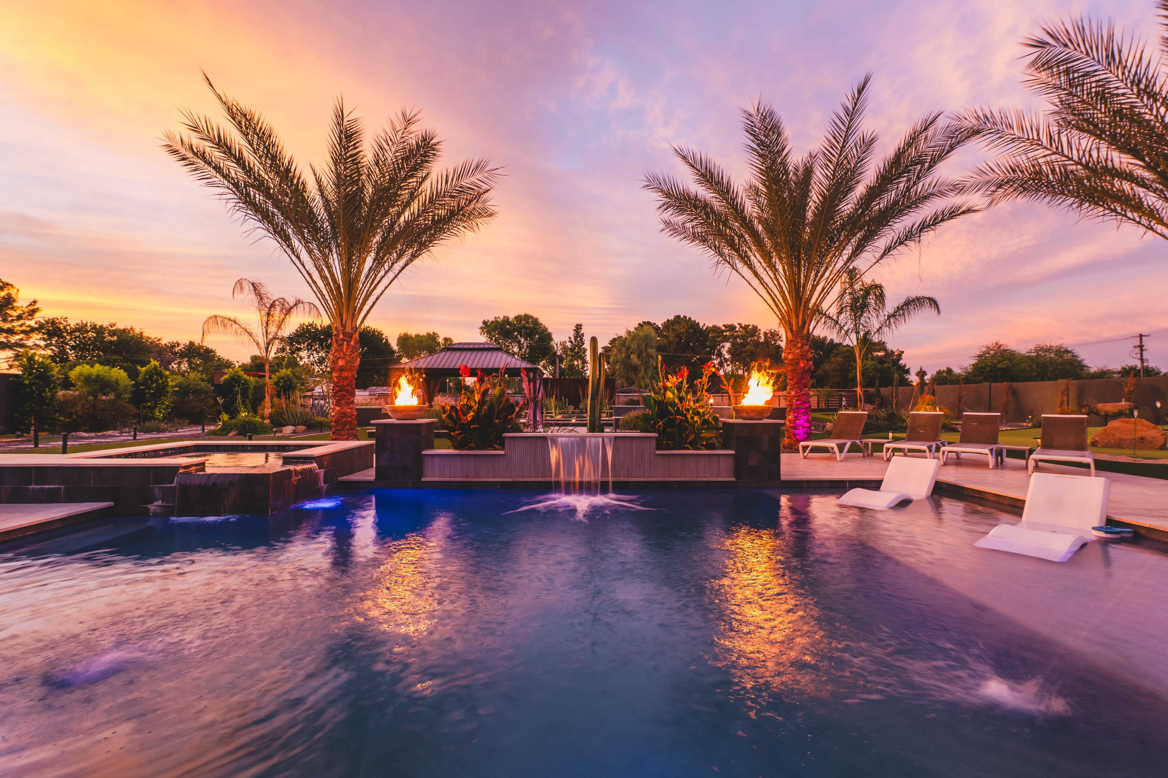 The image shows a backyard pool with palm trees, surrounding landscaping, and illuminated features under a colorful sunset sky.
