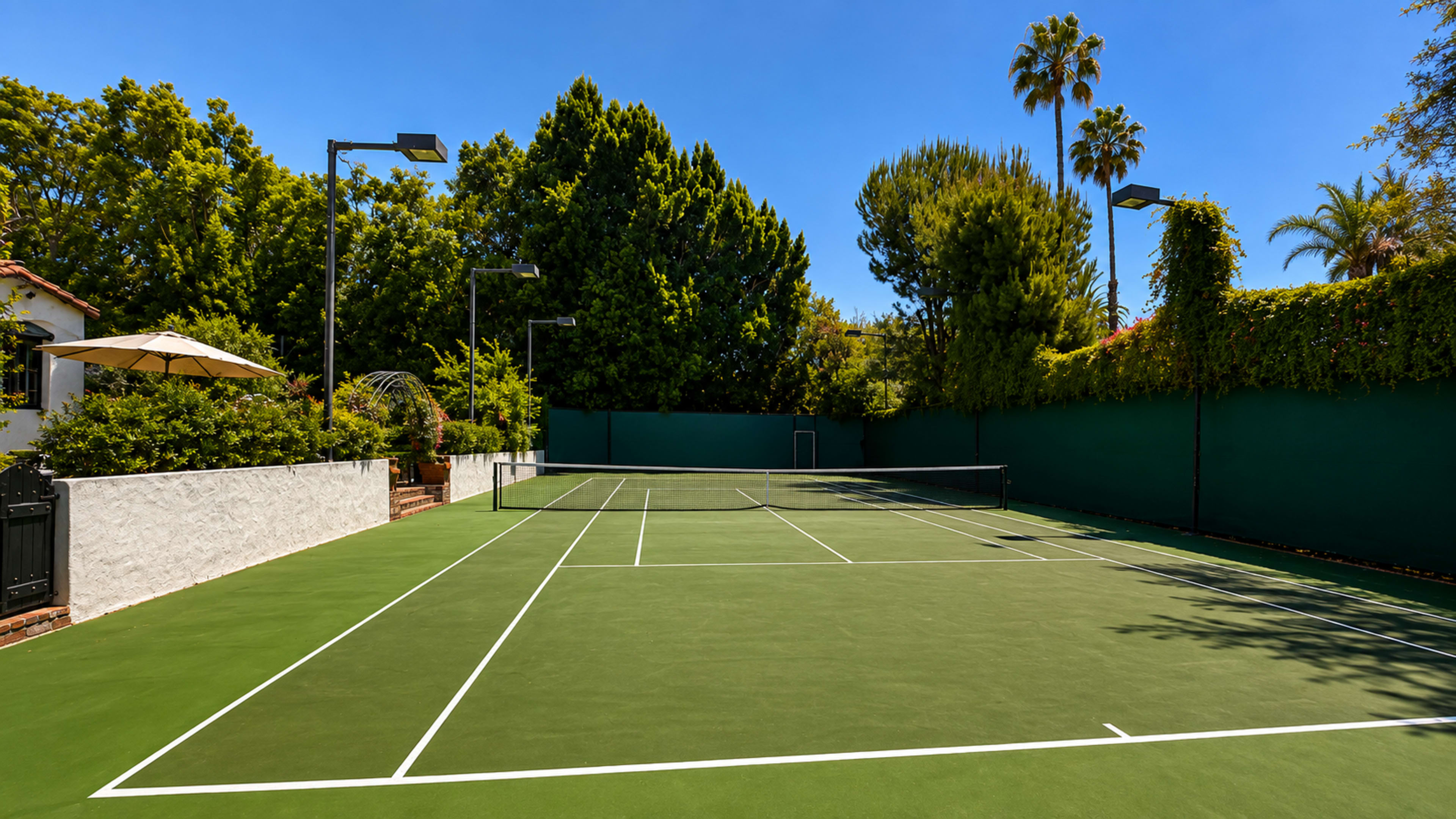 The image shows a well-maintained tennis court surrounded by lush greenery and palm trees under a clear blue sky.