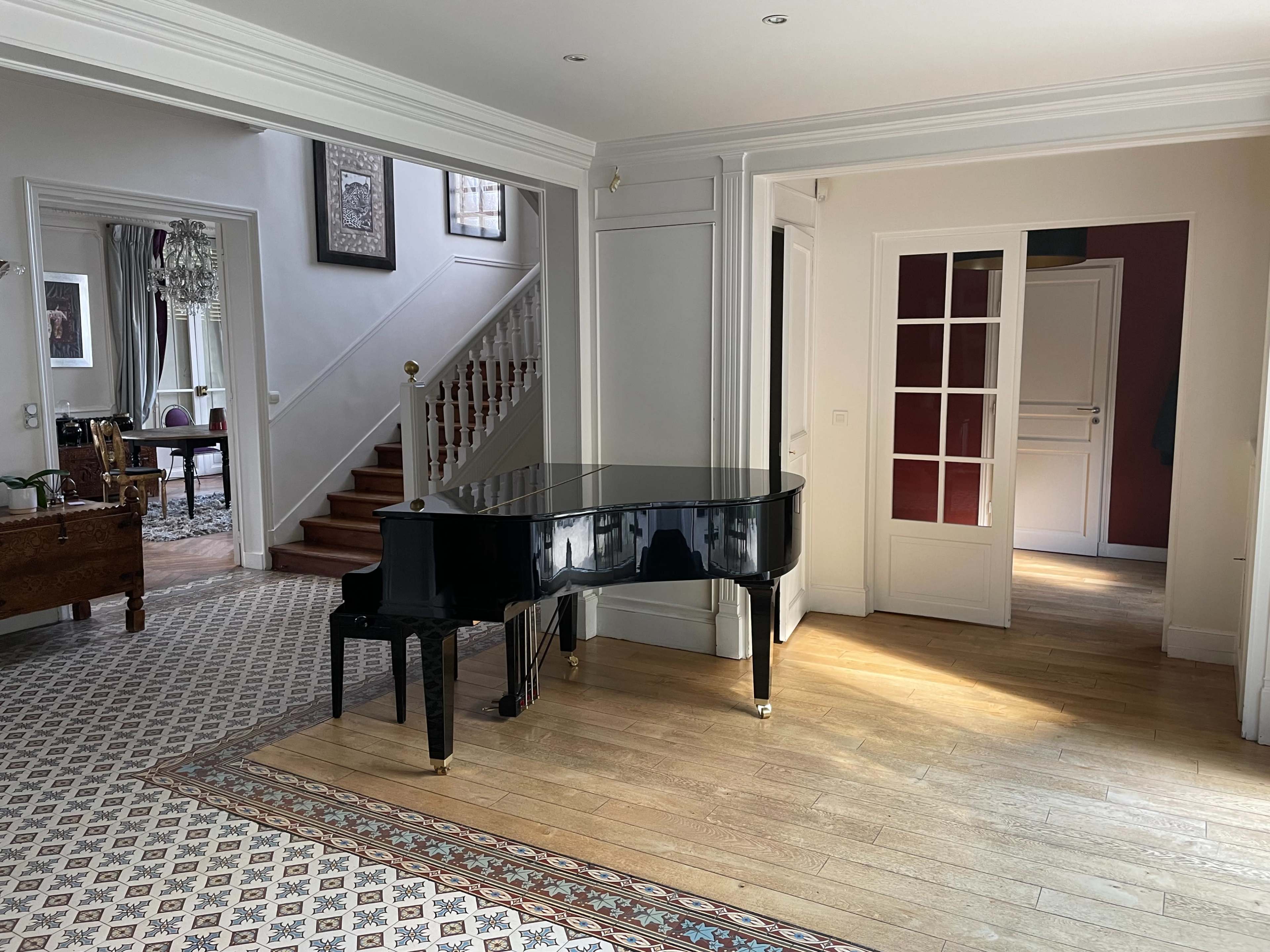 A spacious interior with a grand piano positioned centrally, surrounded by a patterned floor and stairway leading to an upper level.