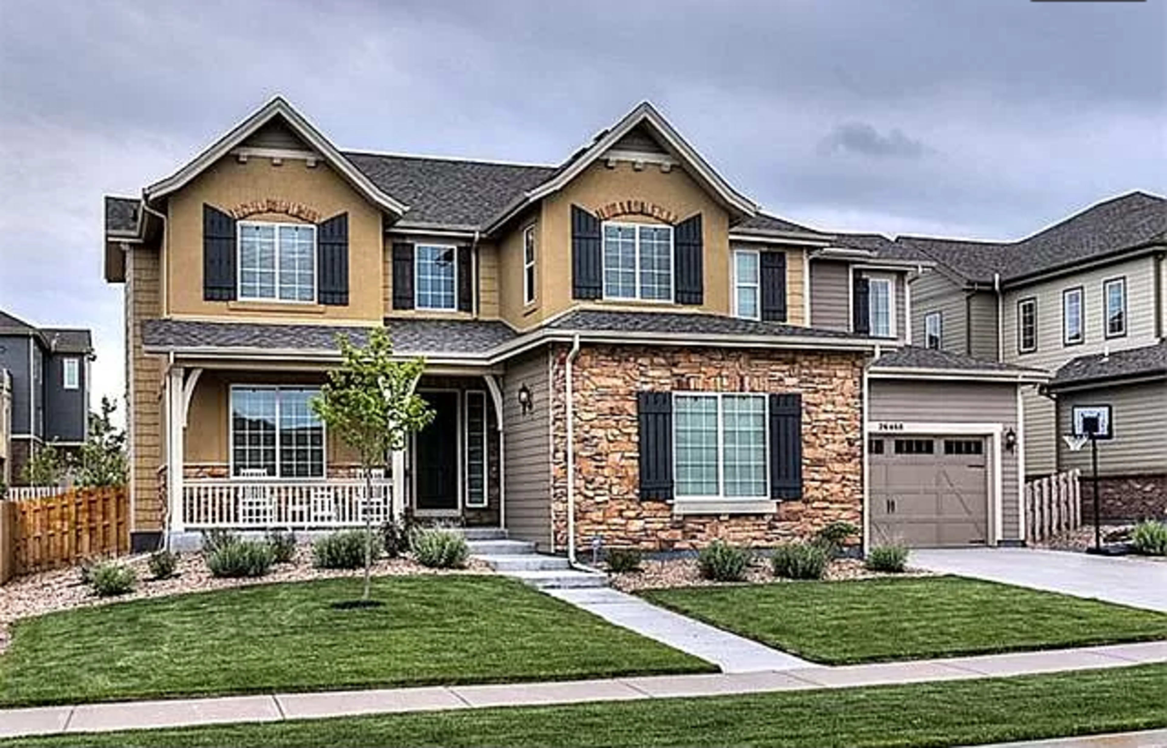 A two-story house features a mix of beige siding and stone with a front porch and a well-manicured lawn.