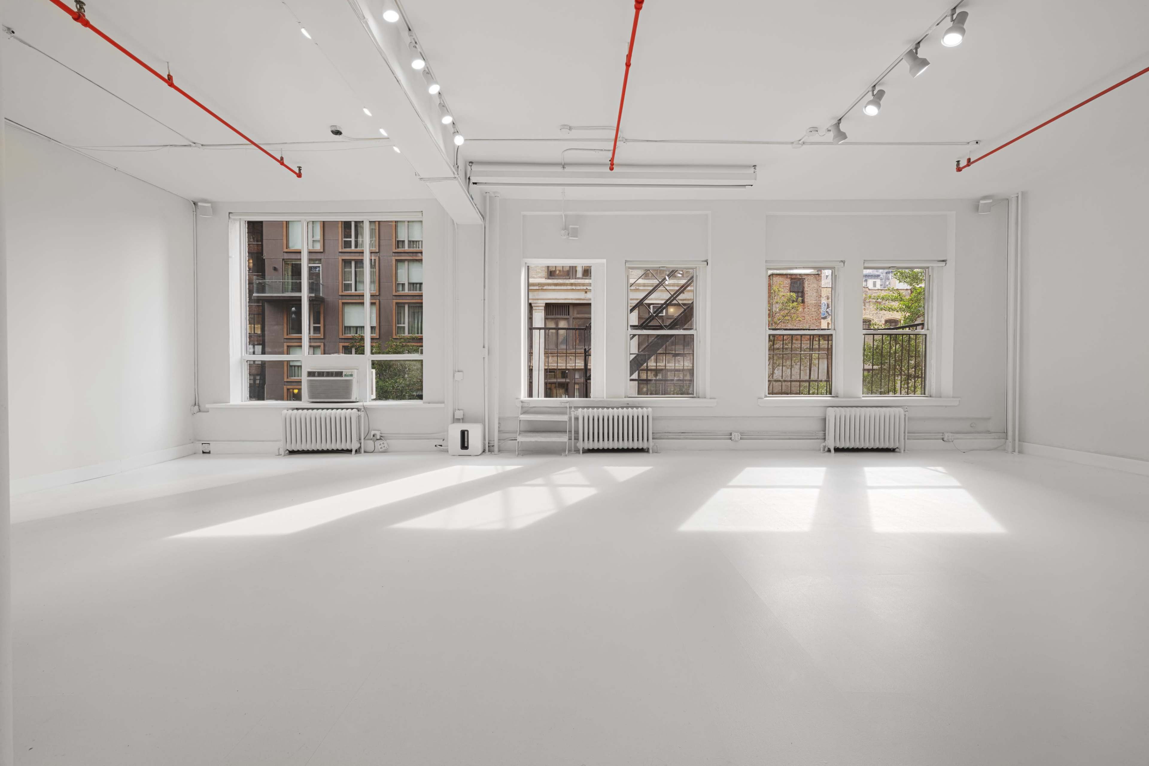 A bright, empty room features white walls and a light-colored floor with large windows letting in natural light.