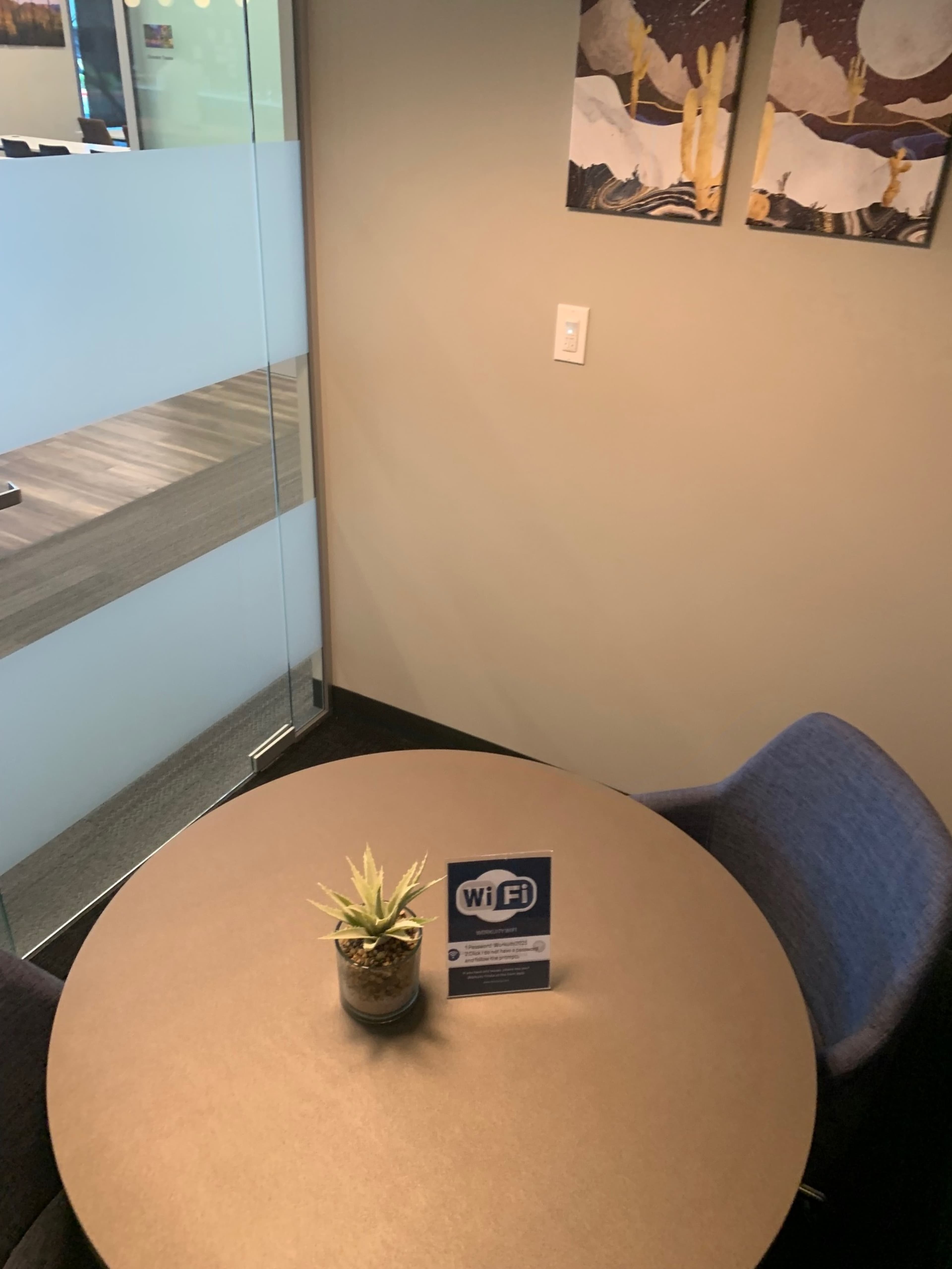 A small round table with a potted plant and a Wi-Fi information sign in a corner of an office with glass walls.