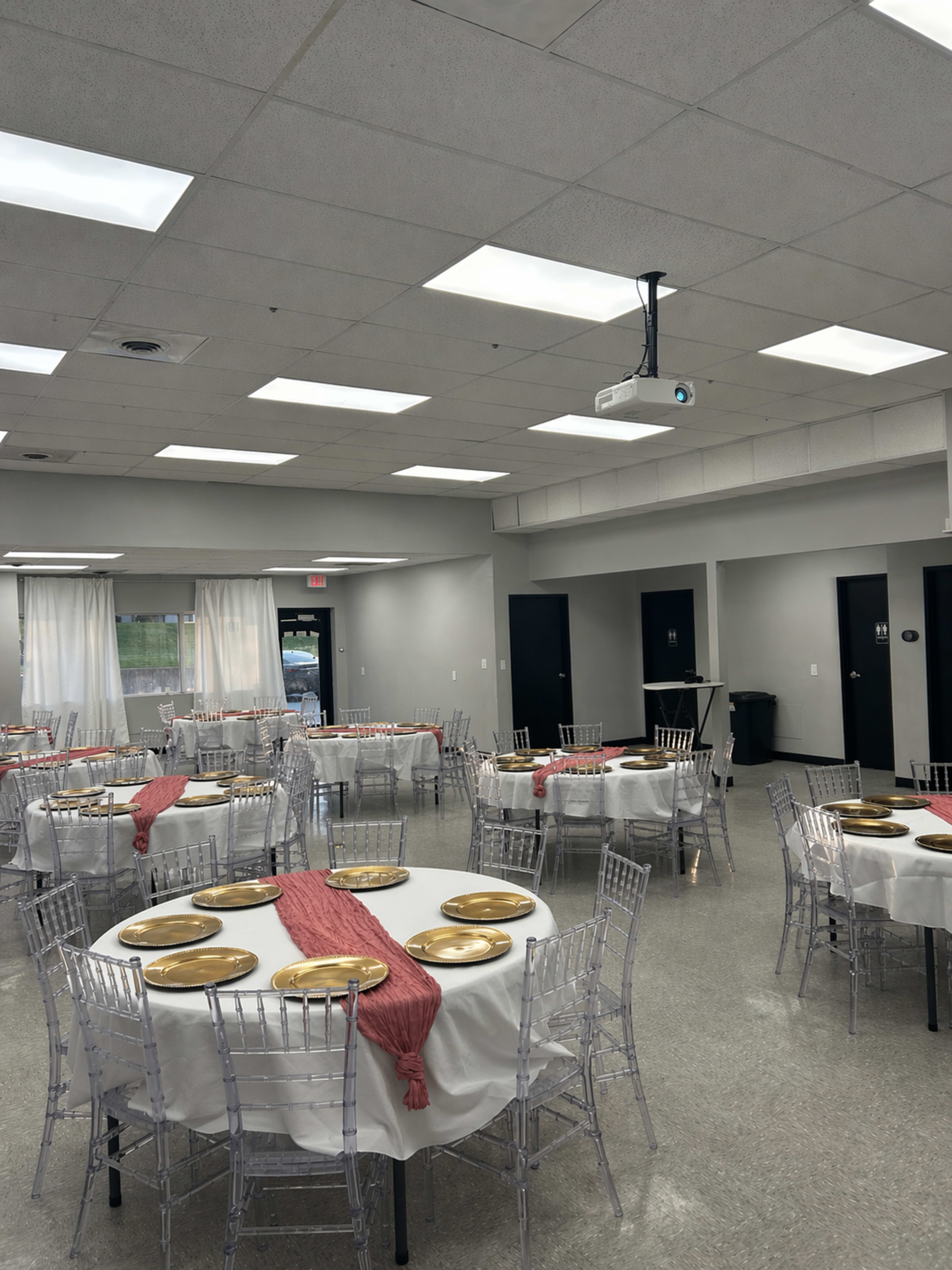 The image shows a well-lit banquet room with several round tables set with white linens, gold plates, and red runners.