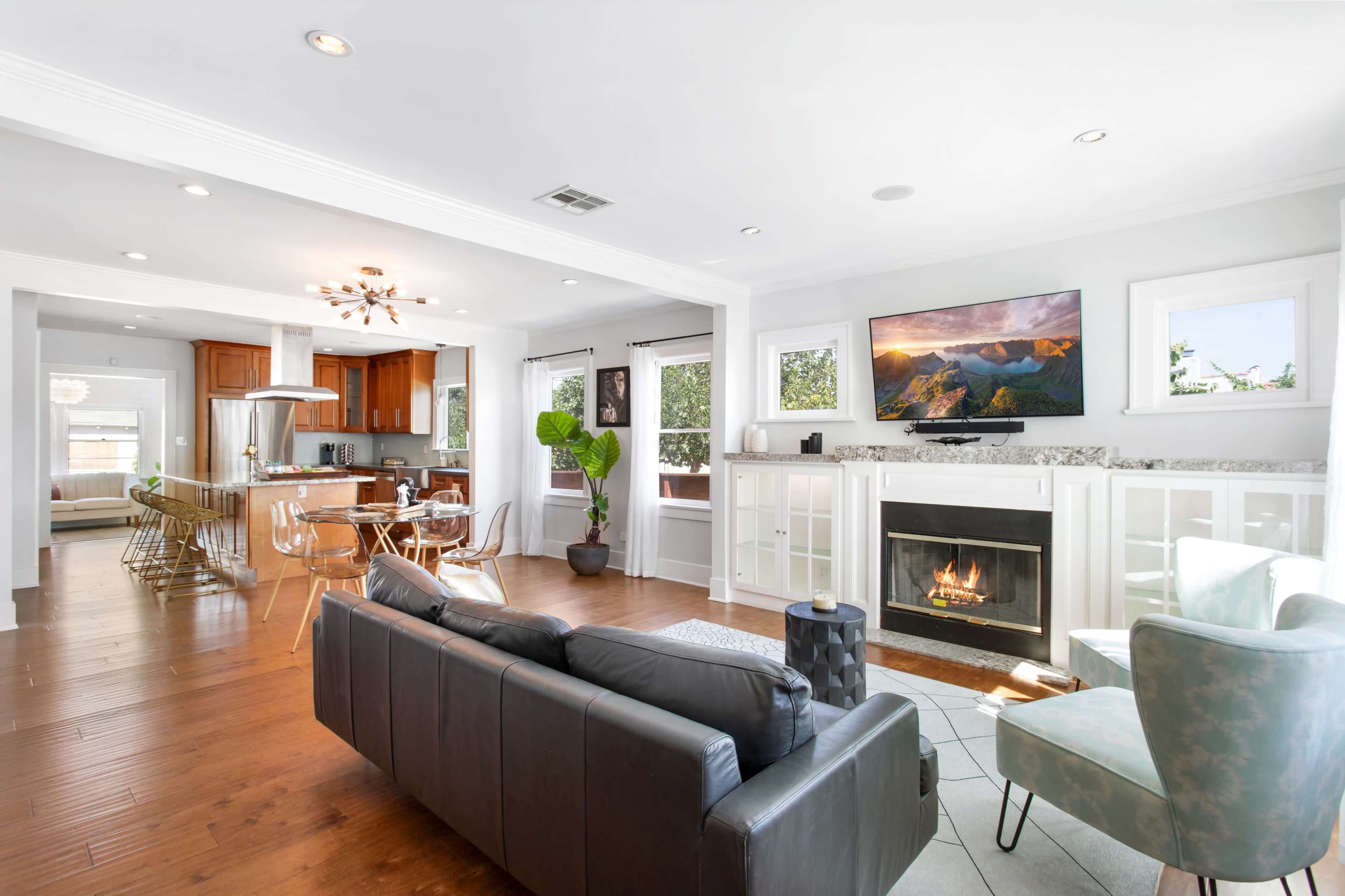 A living room features a black sofa, a light blue armchair, a fireplace with a TV above it, and an open kitchen area with wooden cabinets in the background.