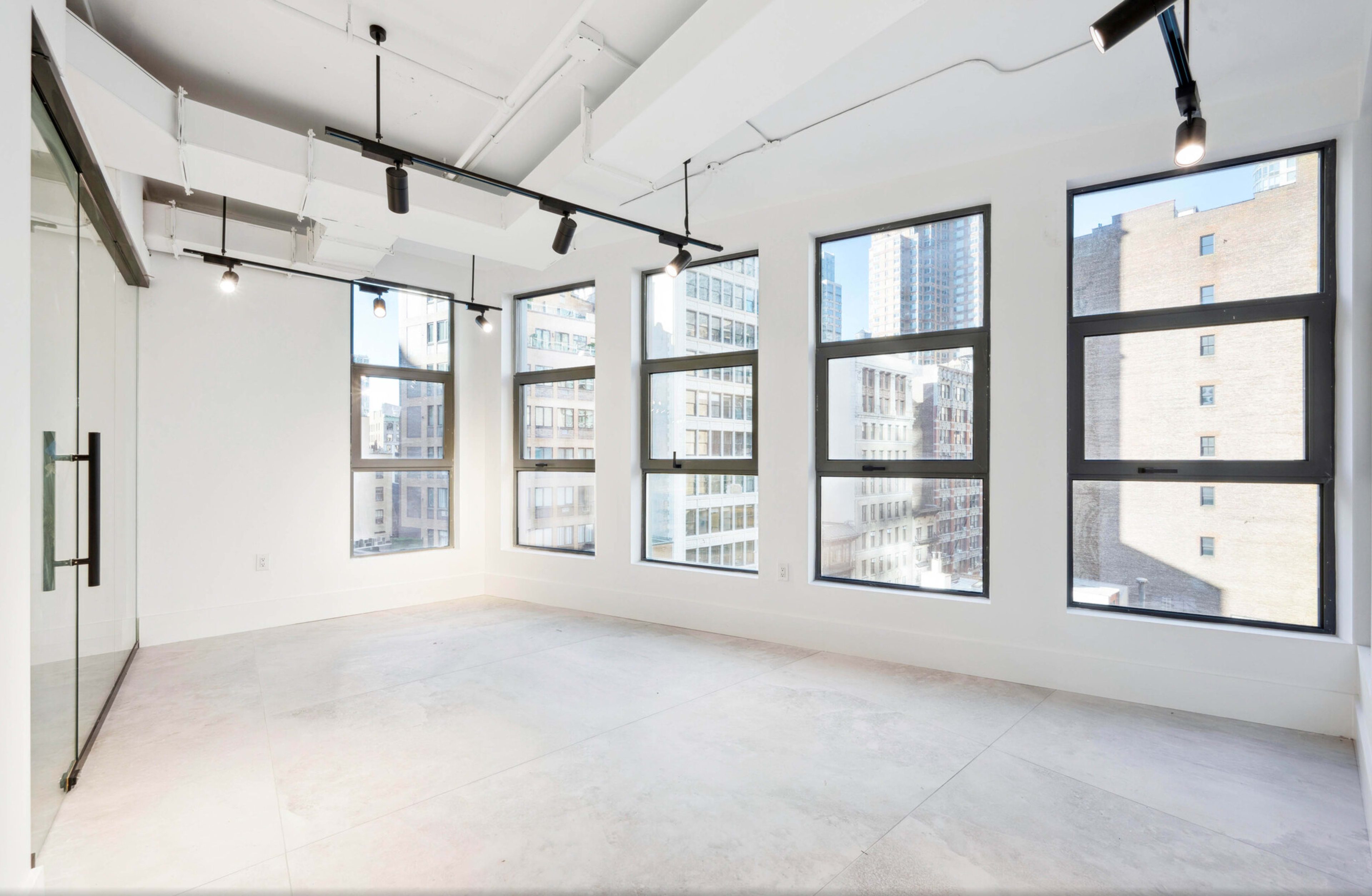 The image shows an empty, well-lit room with large windows on two sides and a glass-walled partition.