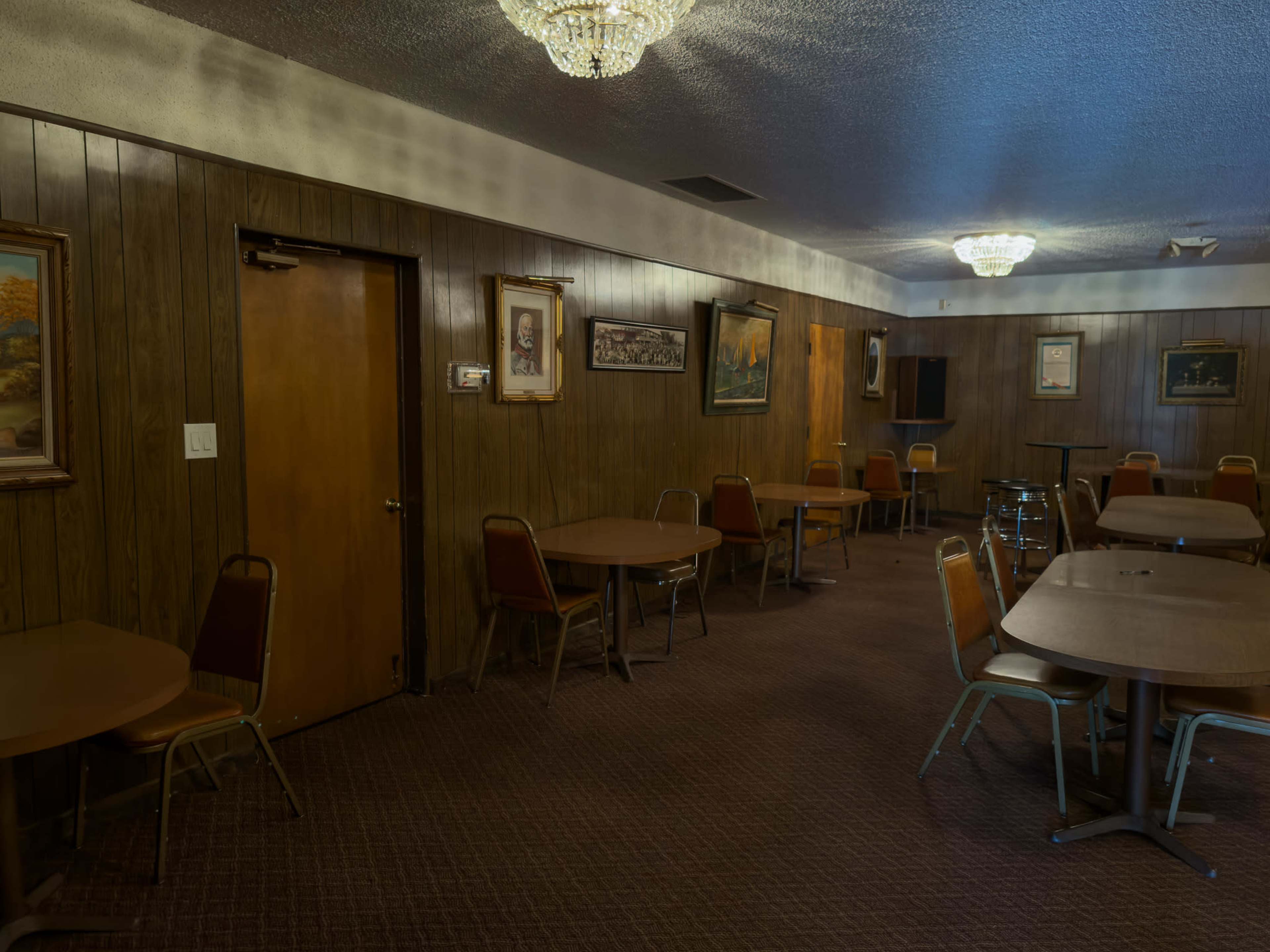 The image shows a dimly lit room with wooden paneling, featuring several empty tables and chairs, along with wall-mounted art and a door on one side.