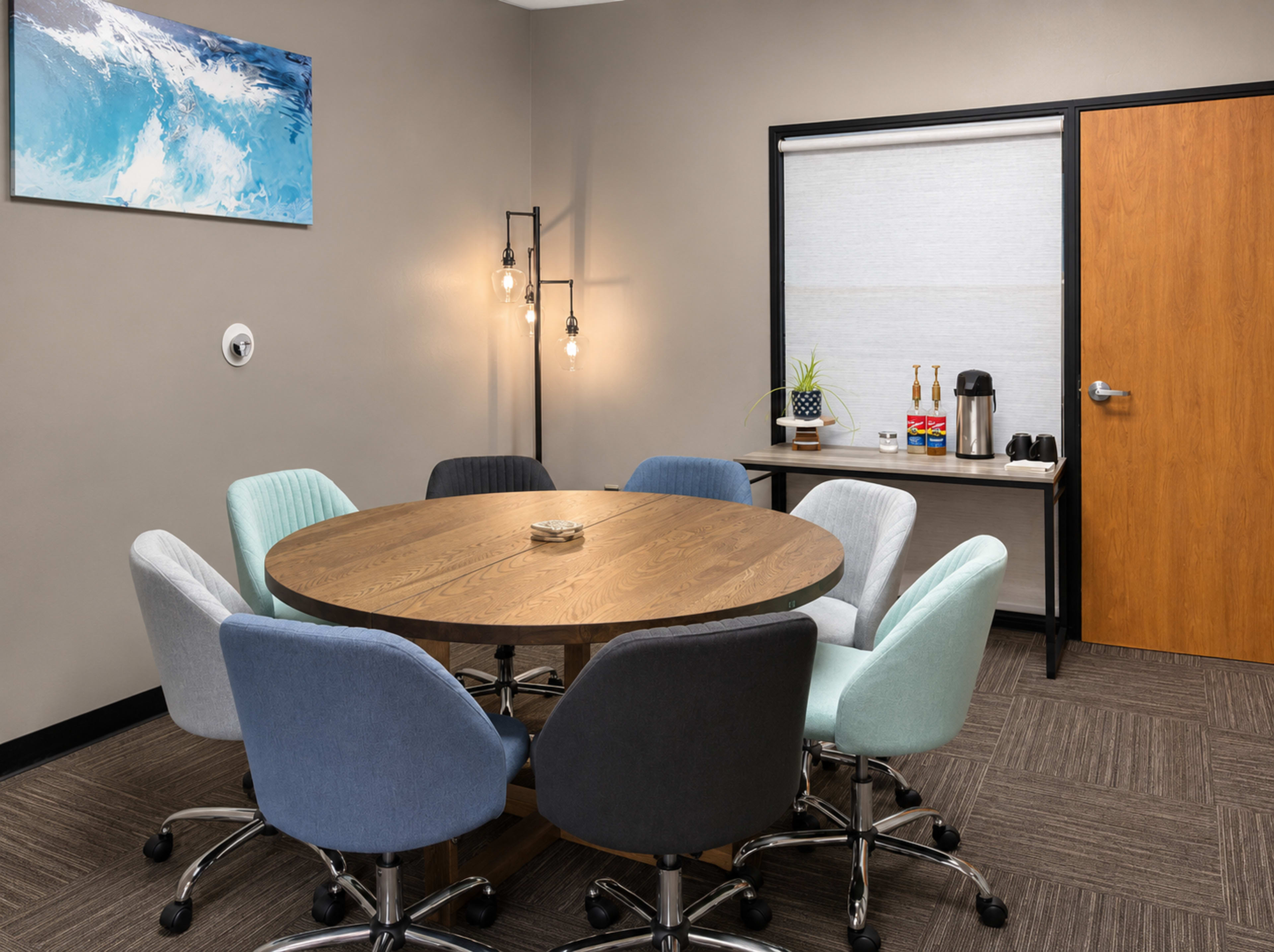 A round wooden table surrounded by eight chairs in a well-lit conference room, with a kitchenette featuring a coffee machine and refreshments nearby.