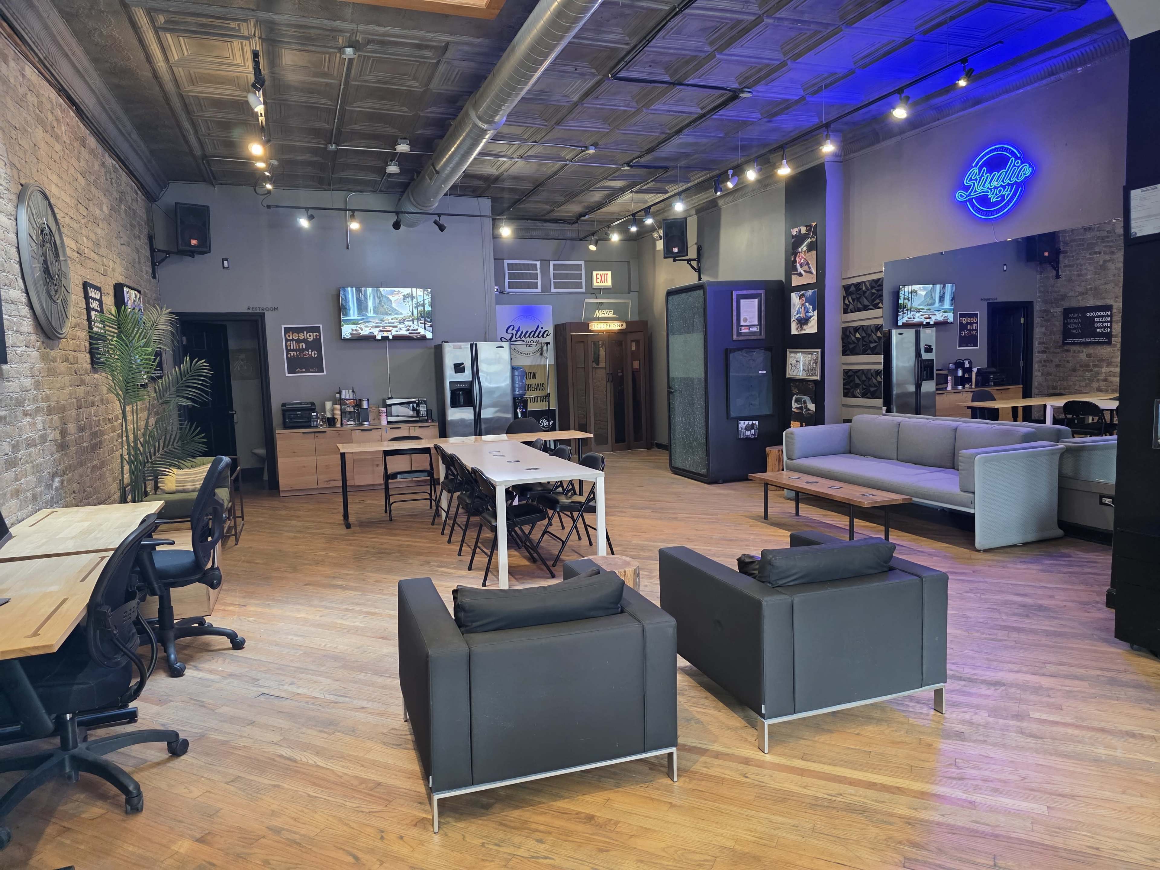 The image shows a modern space with wooden flooring, featuring a mix of seating areas including black couches, dining tables, and screens mounted on the walls.