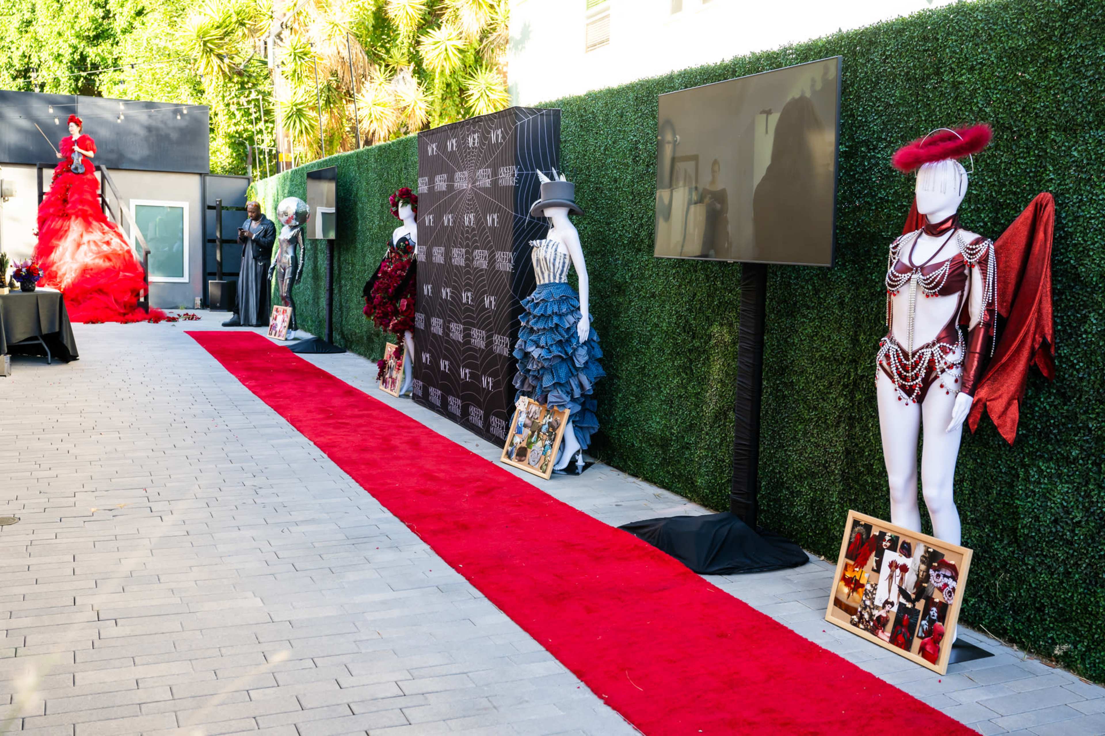 A red carpet event with decorative mannequins dressed in elaborate costumes lined against a green hedged backdrop.