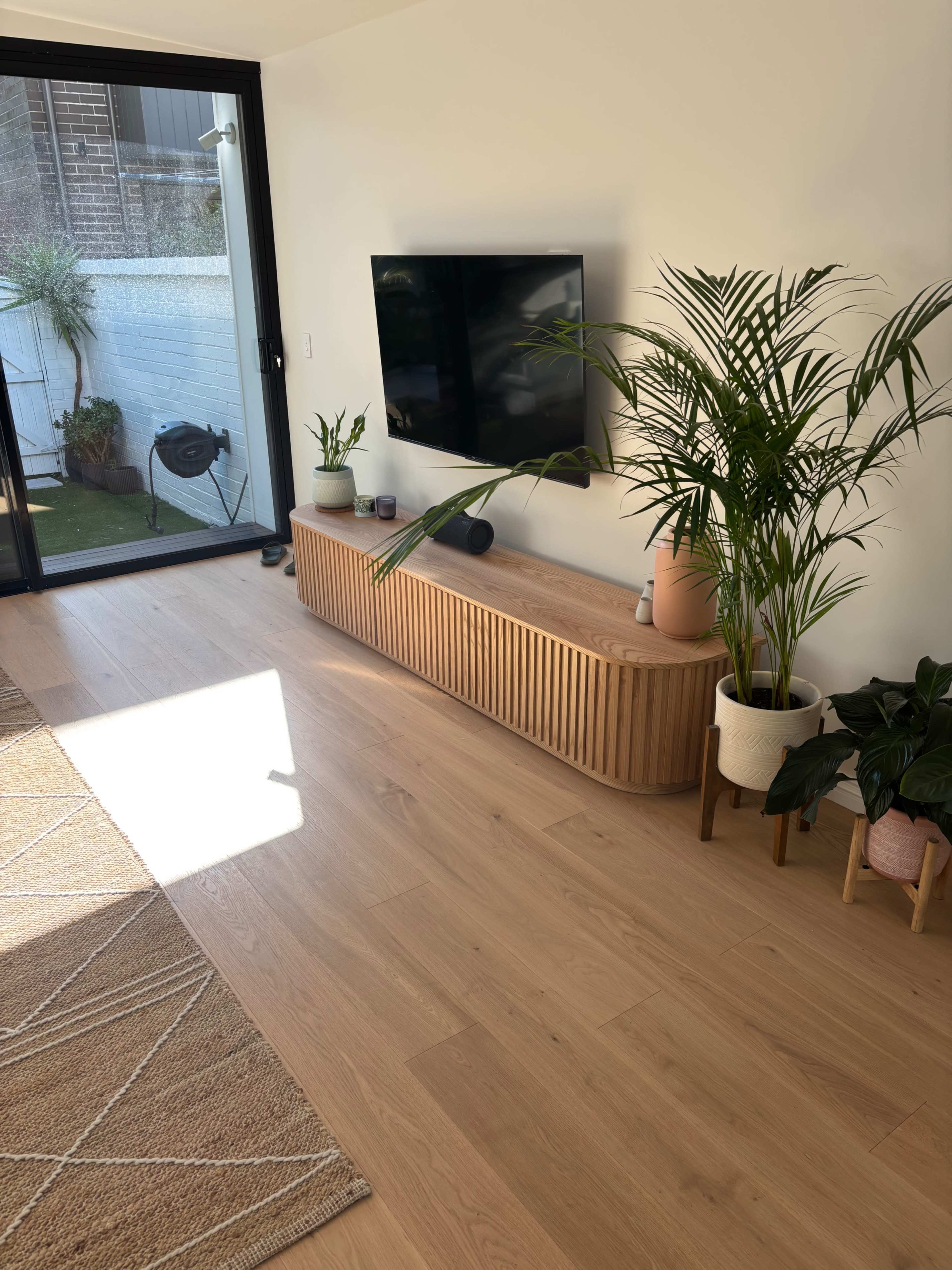 The image shows a modern living room with a wooden TV stand, indoor plants, and large windows allowing natural light.