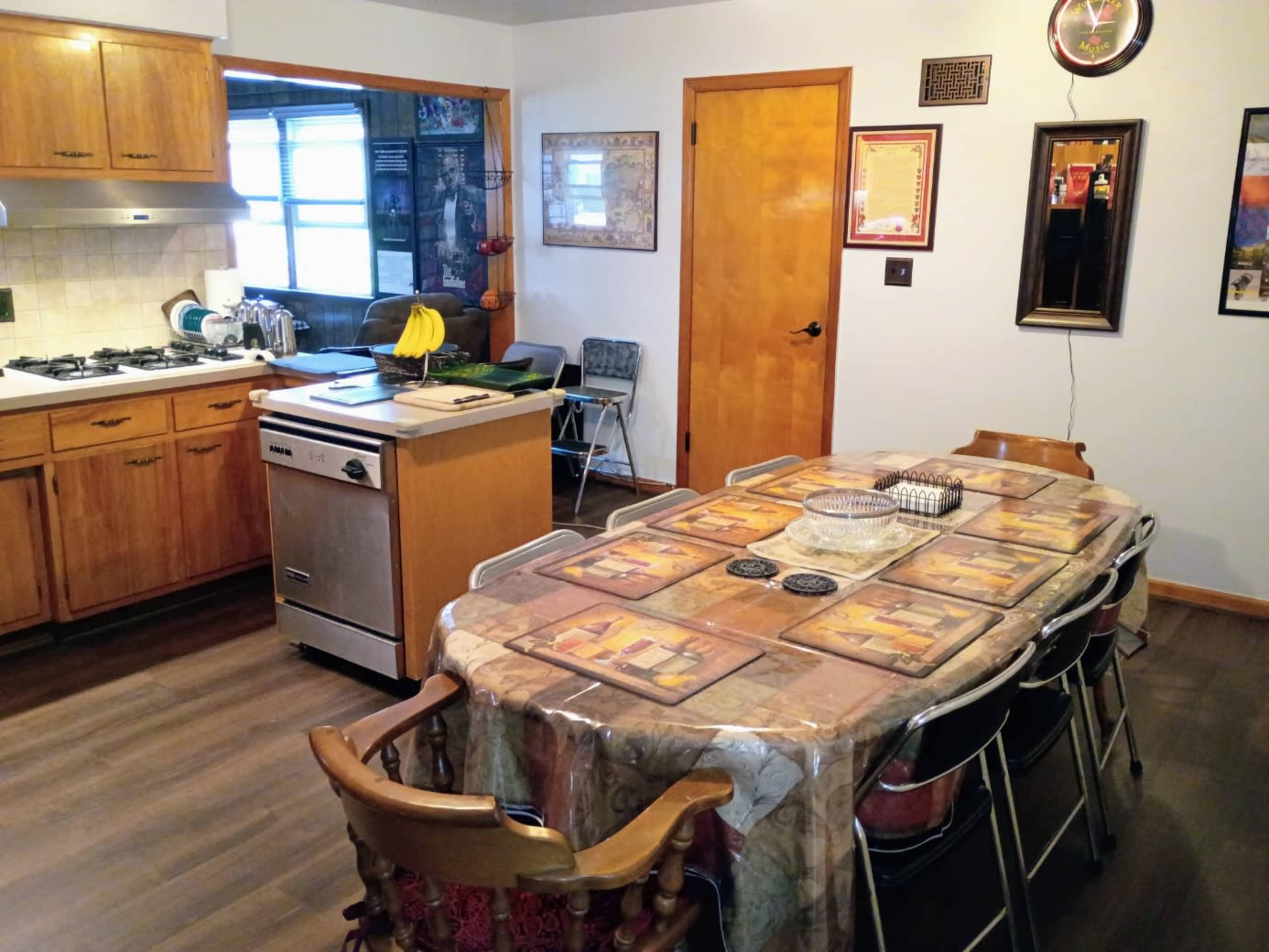 The image shows a kitchen and dining area featuring wooden cabinetry, a dining table with place mats, and a kitchen island with a dishwasher in the background.