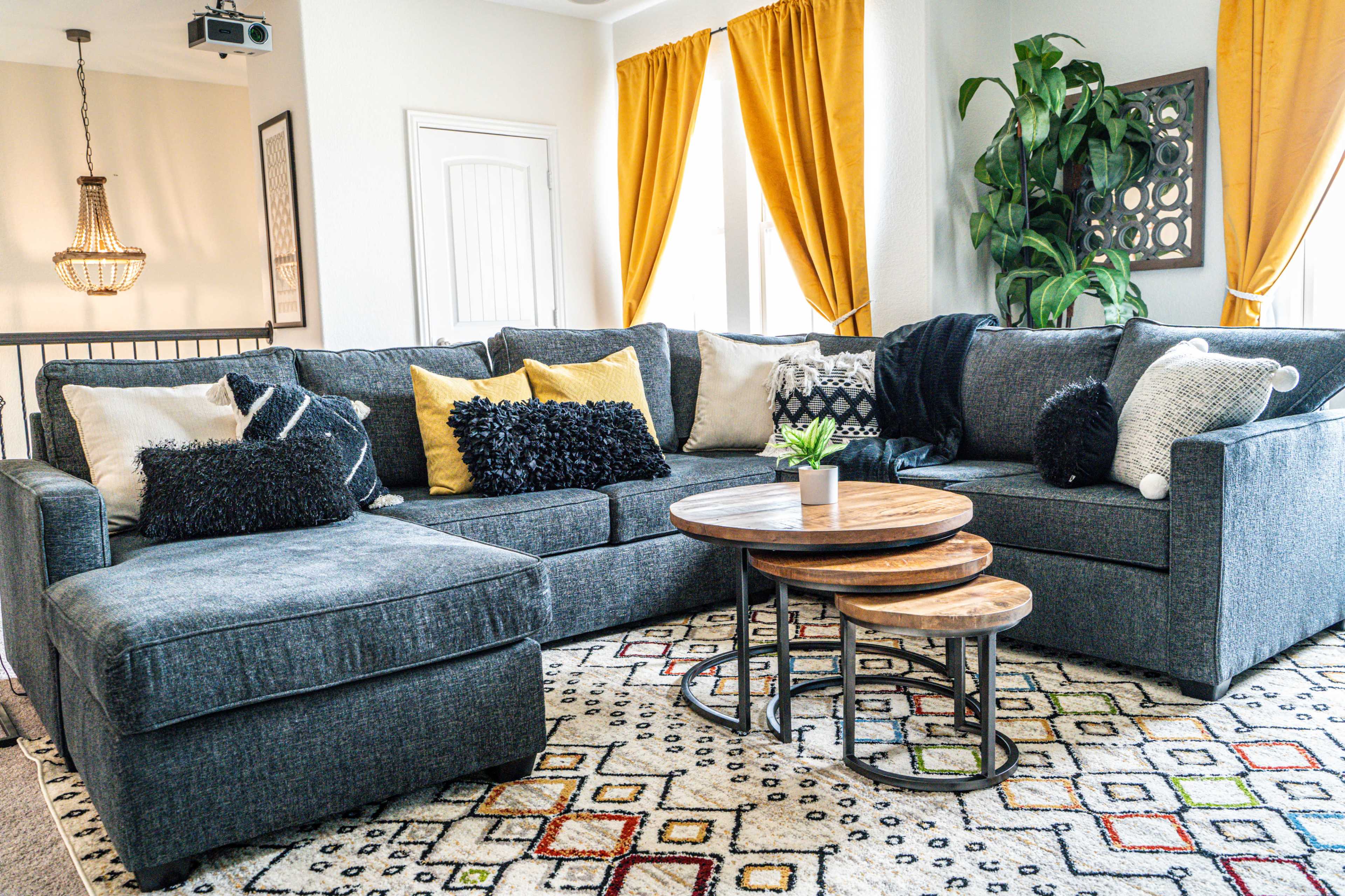 A modern living room features a large gray sectional sofa, two wooden coffee tables, a patterned rug, and bright yellow curtains.