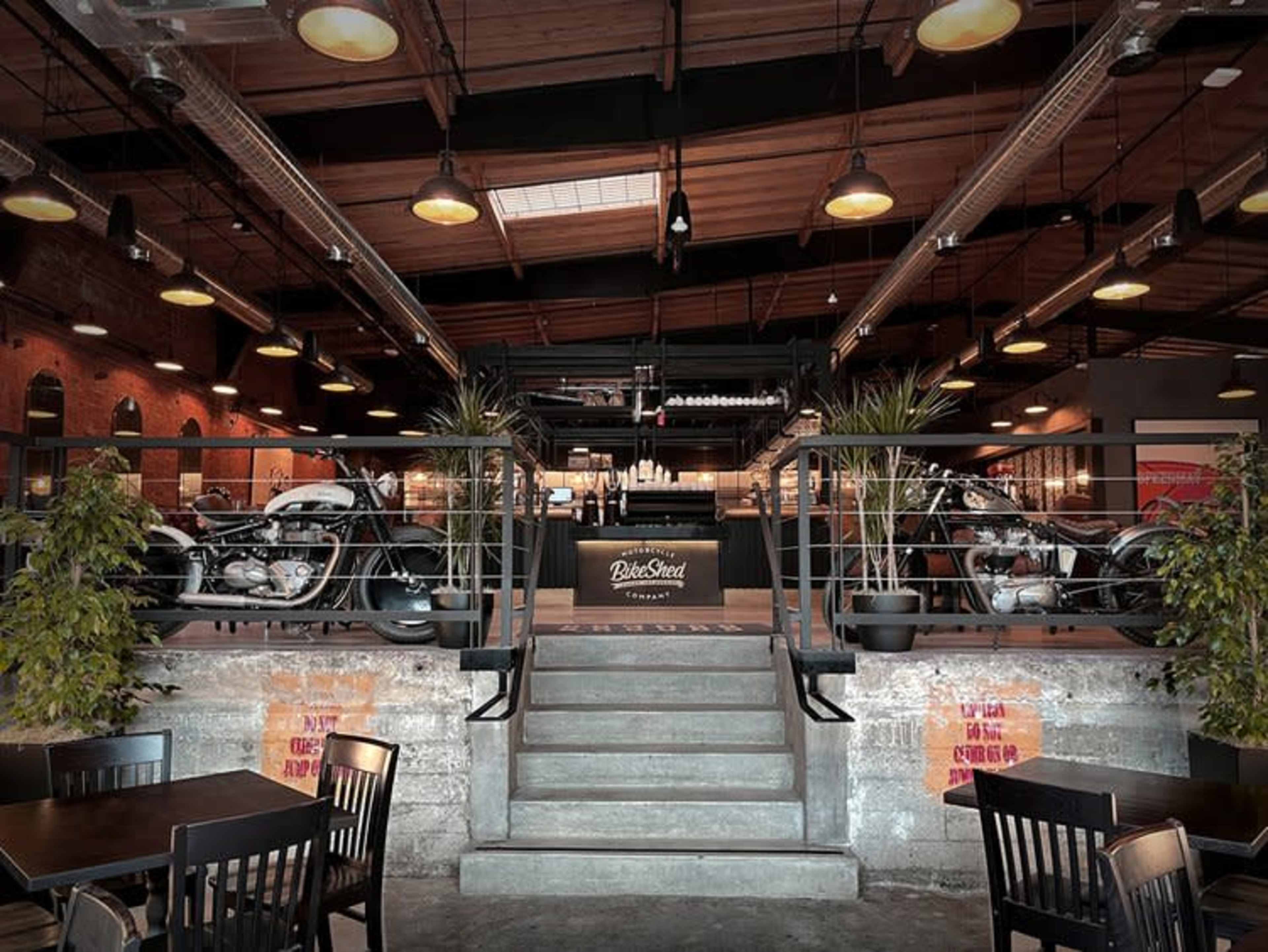 The image shows a modern restaurant interior featuring a staircase leading to an upper level, with motorcycle displays on either side and dark wooden furnishings.