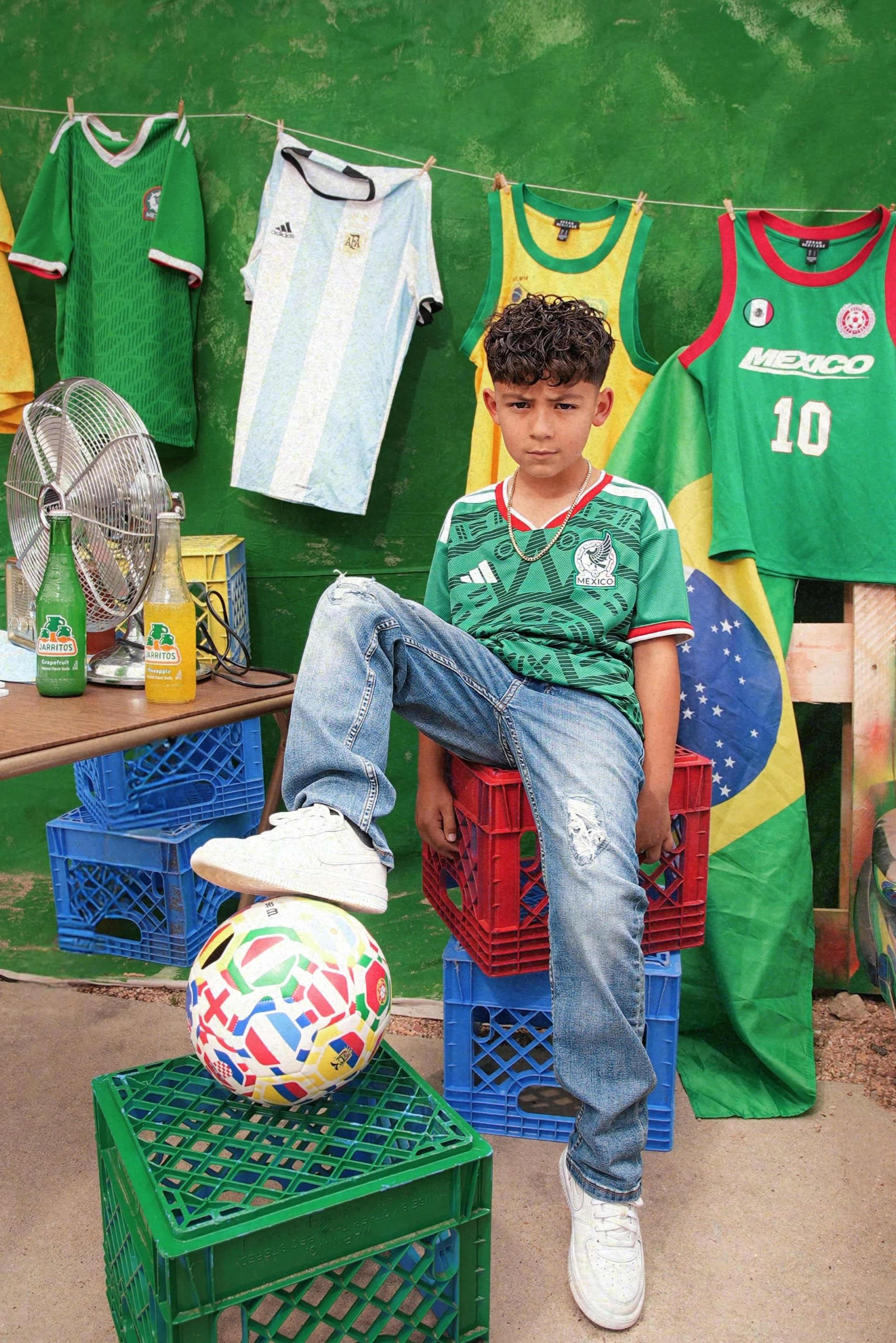 A boy wearing a green soccer jersey sits on a stack of crates with a colorful soccer ball at his feet, surrounded by soccer jerseys and flags hanging on a green wall.
