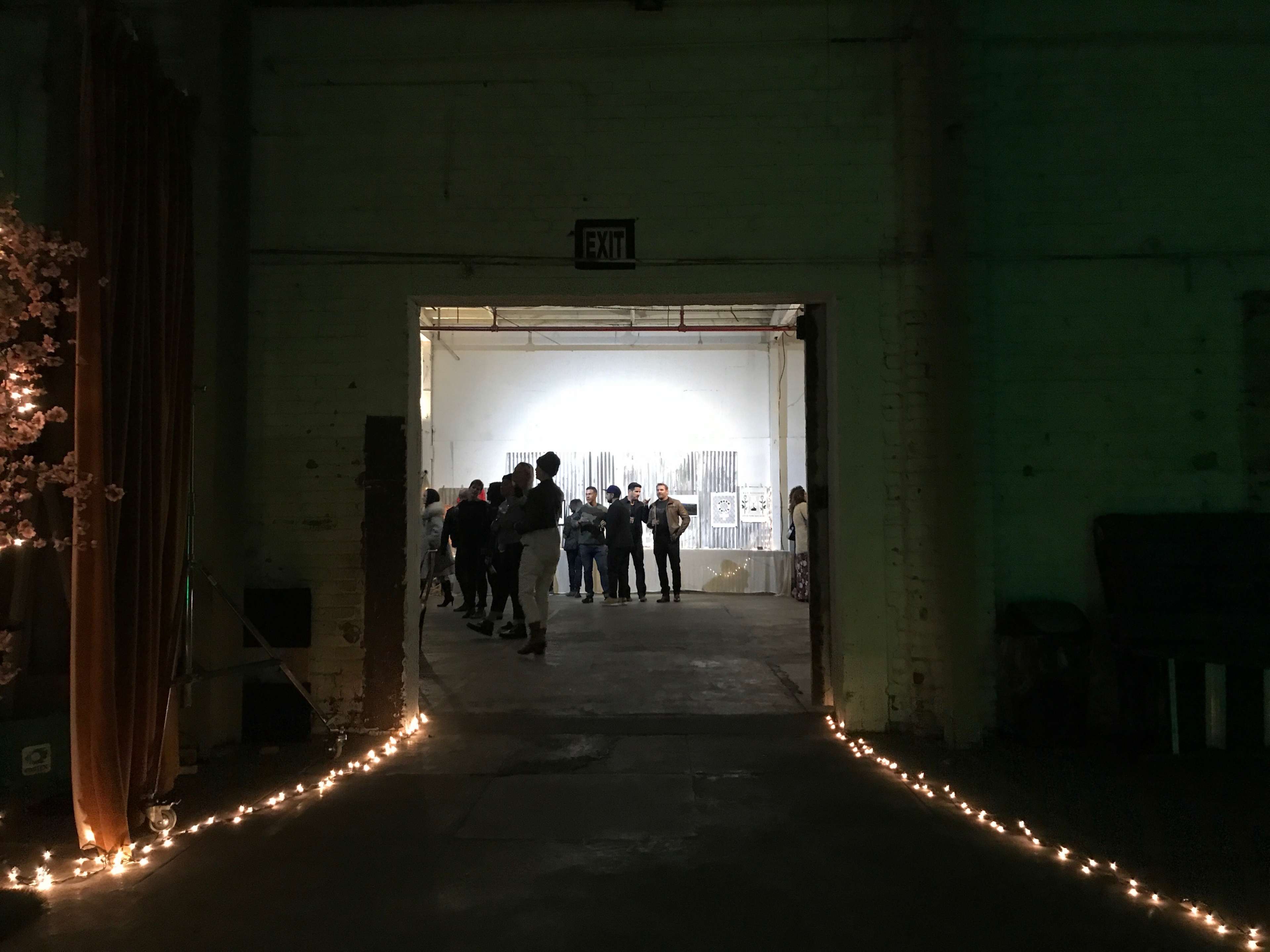 People are gathered in an industrial space, visible through an open doorway illuminated by string lights along the floor.