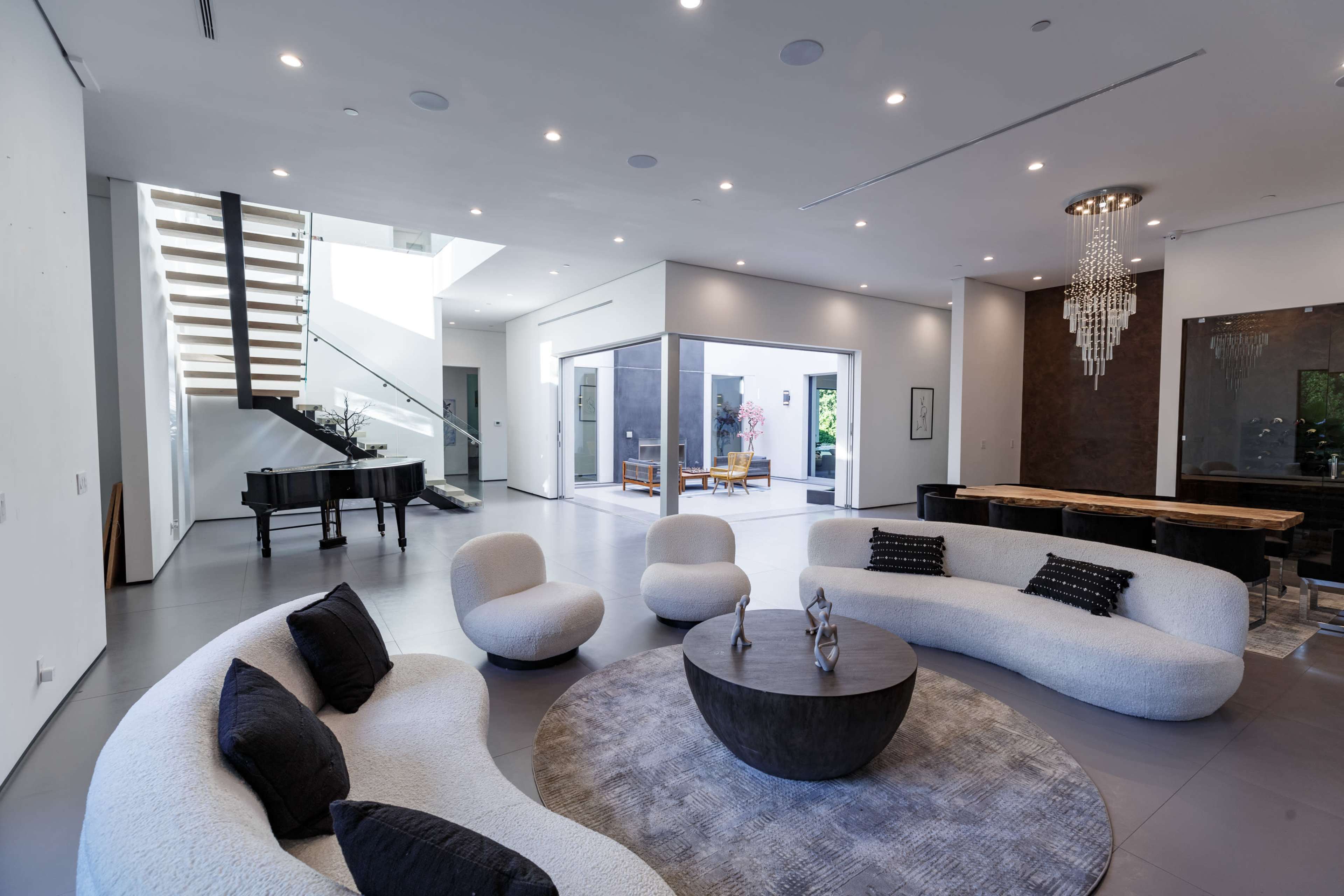 The image shows a modern living room featuring a curved white sofa, a round coffee table, a grand piano, and a staircase leading to an upper level, with large windows allowing natural light.