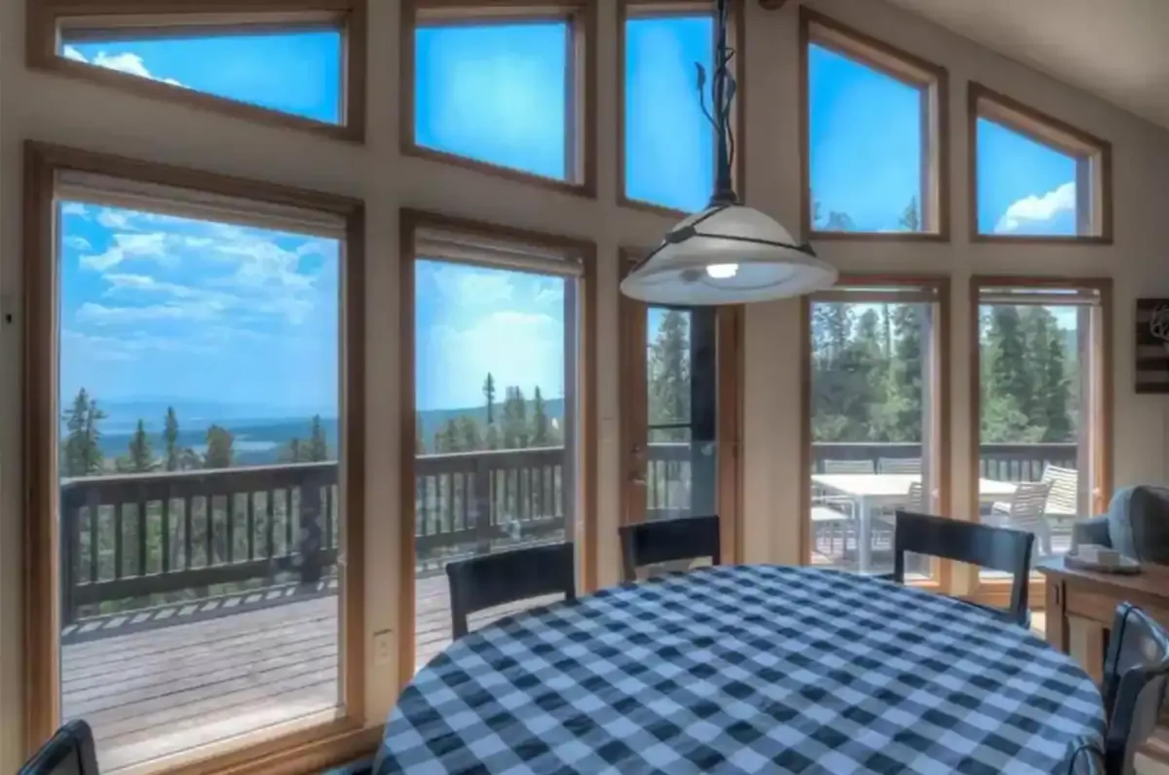 A dining area with a round checkered table and large windows overlooking a wooded landscape and blue sky.