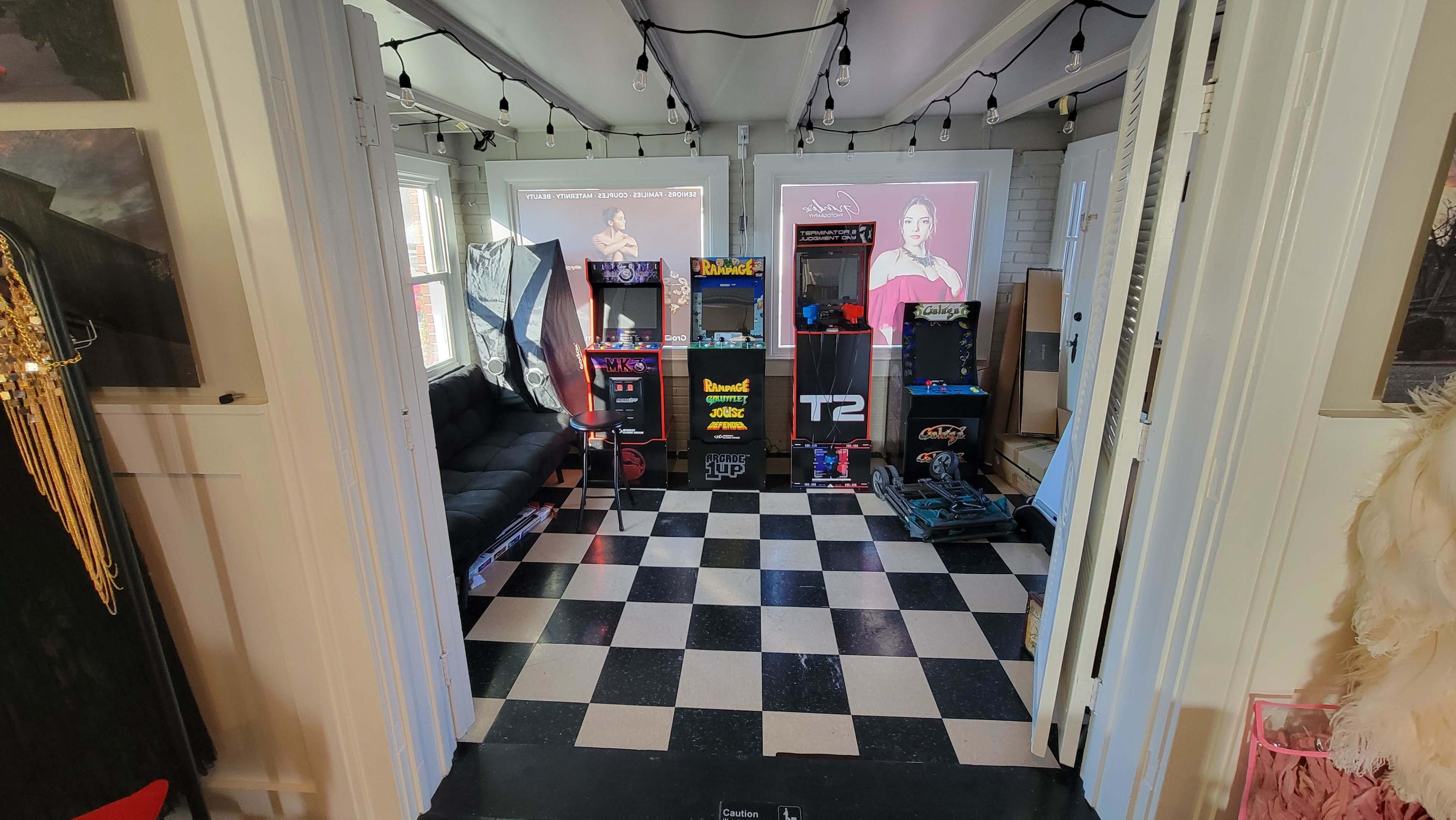 A game room with a checkered floor, displaying rows of arcade machines and a black couch, illuminated by string lights.