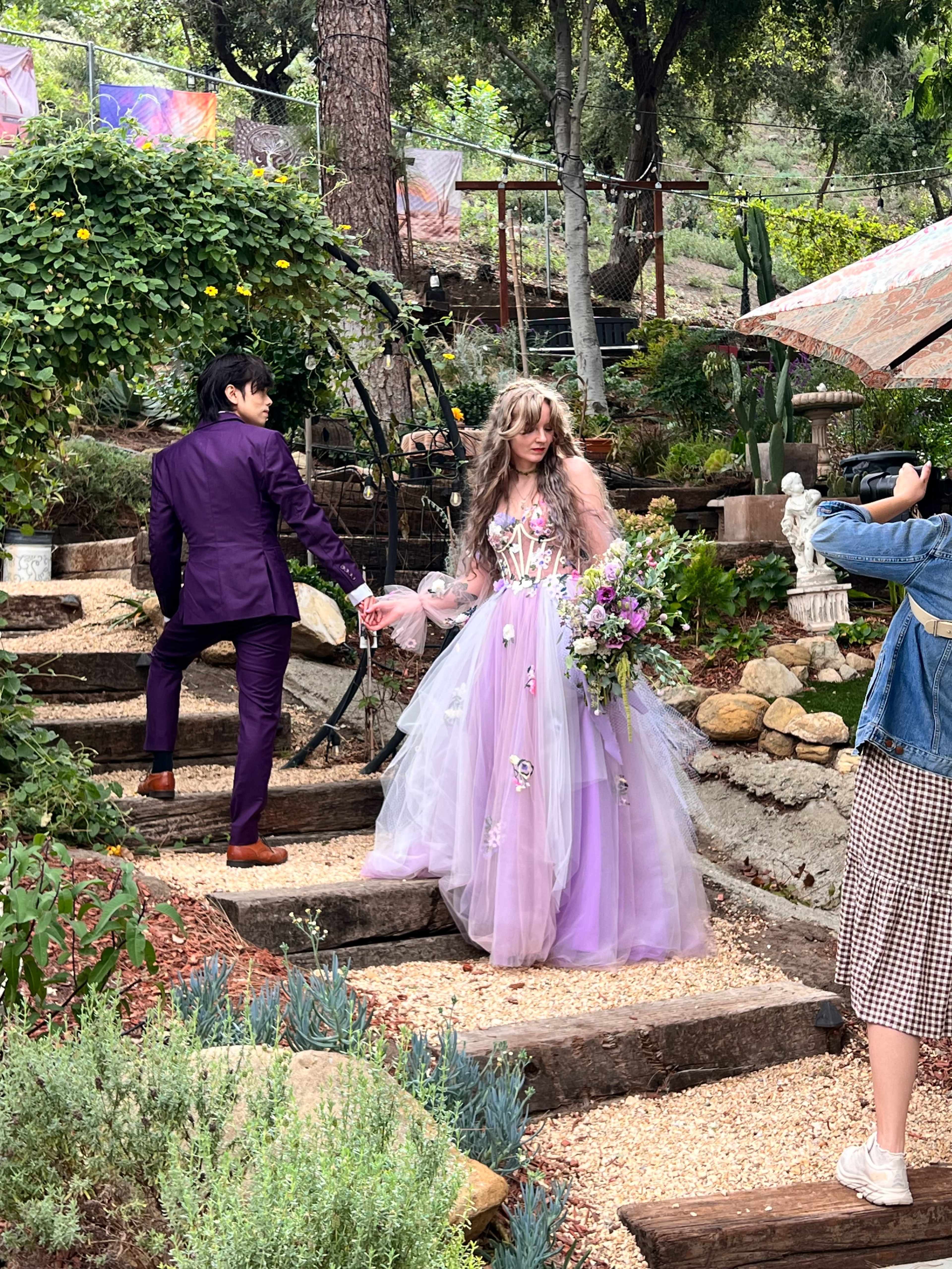 A couple dressed in formal attire poses on a pathway surrounded by greenery and decorative elements, while a person takes a photo of them.