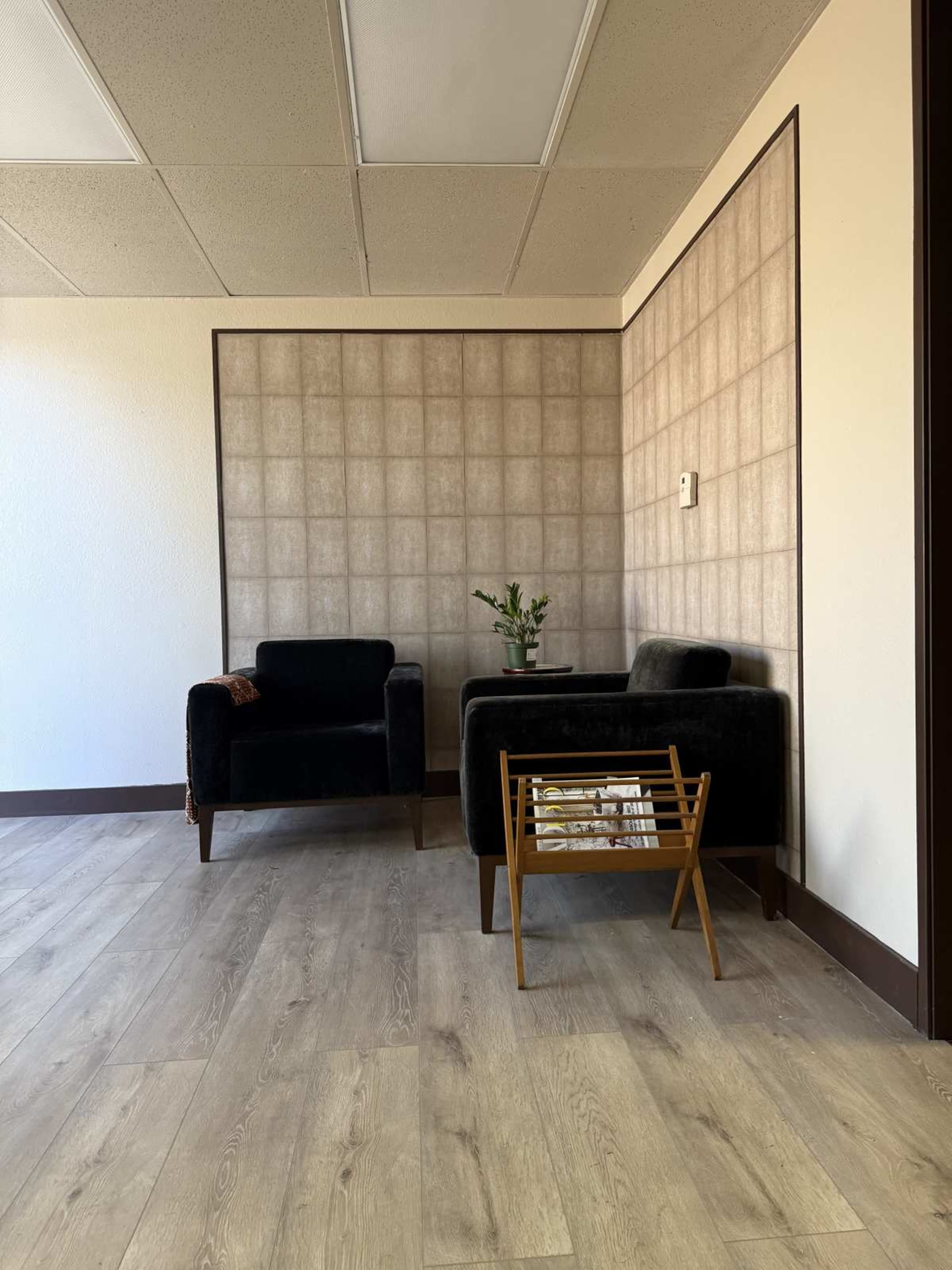 The image shows a simple office corner with two black armchairs, a small side table, and a plant against a textured wall, all on wooden flooring.