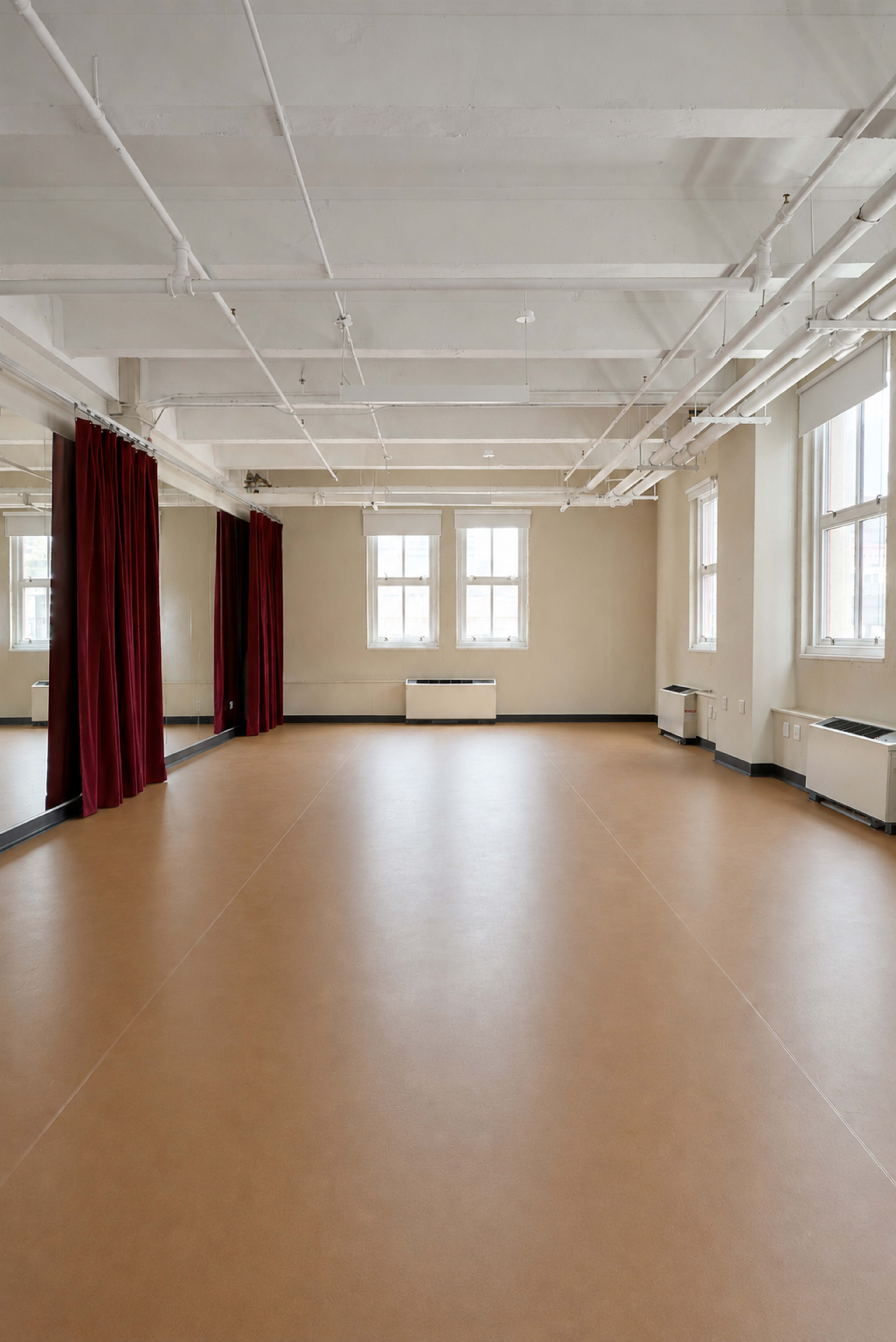 The image shows a spacious, empty room with brown flooring, large windows, and red curtains on two walls.