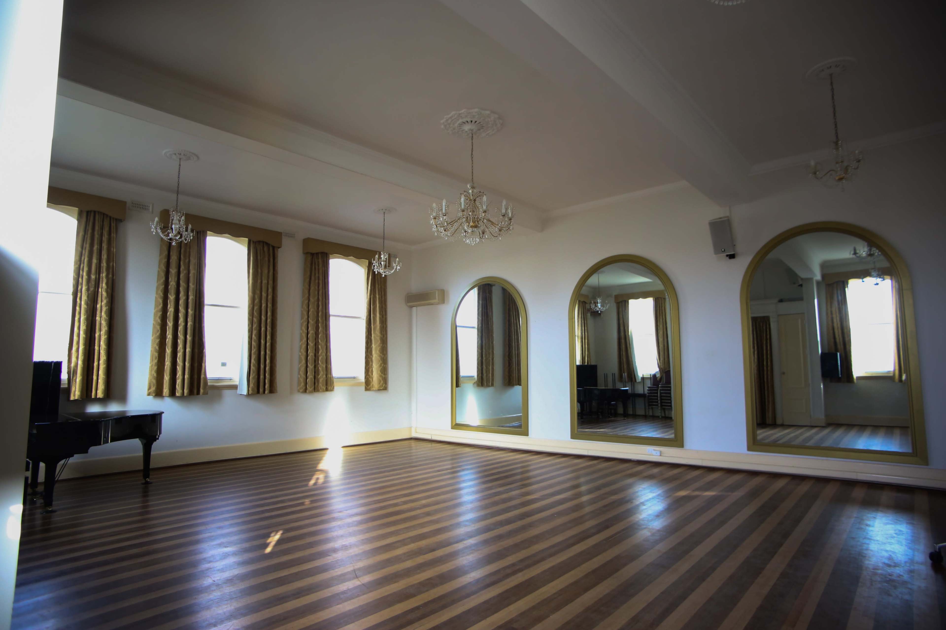 A spacious dance studio with wooden striped flooring, large mirrors, and elegant chandeliers.