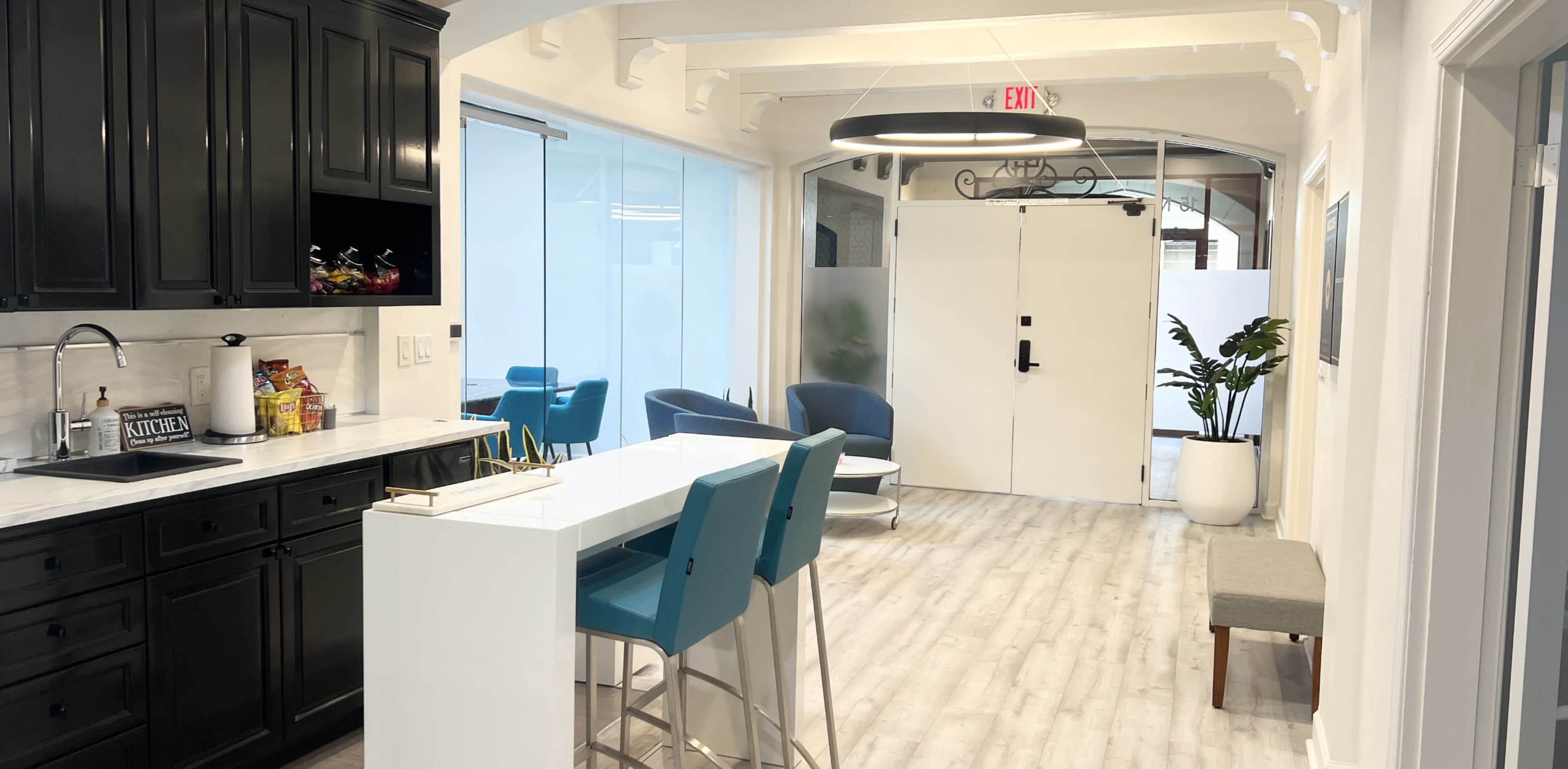 A modern office interior featuring a kitchenette with dark cabinetry, a white island with blue stools, and a lounge area with blue chairs and potted plants.