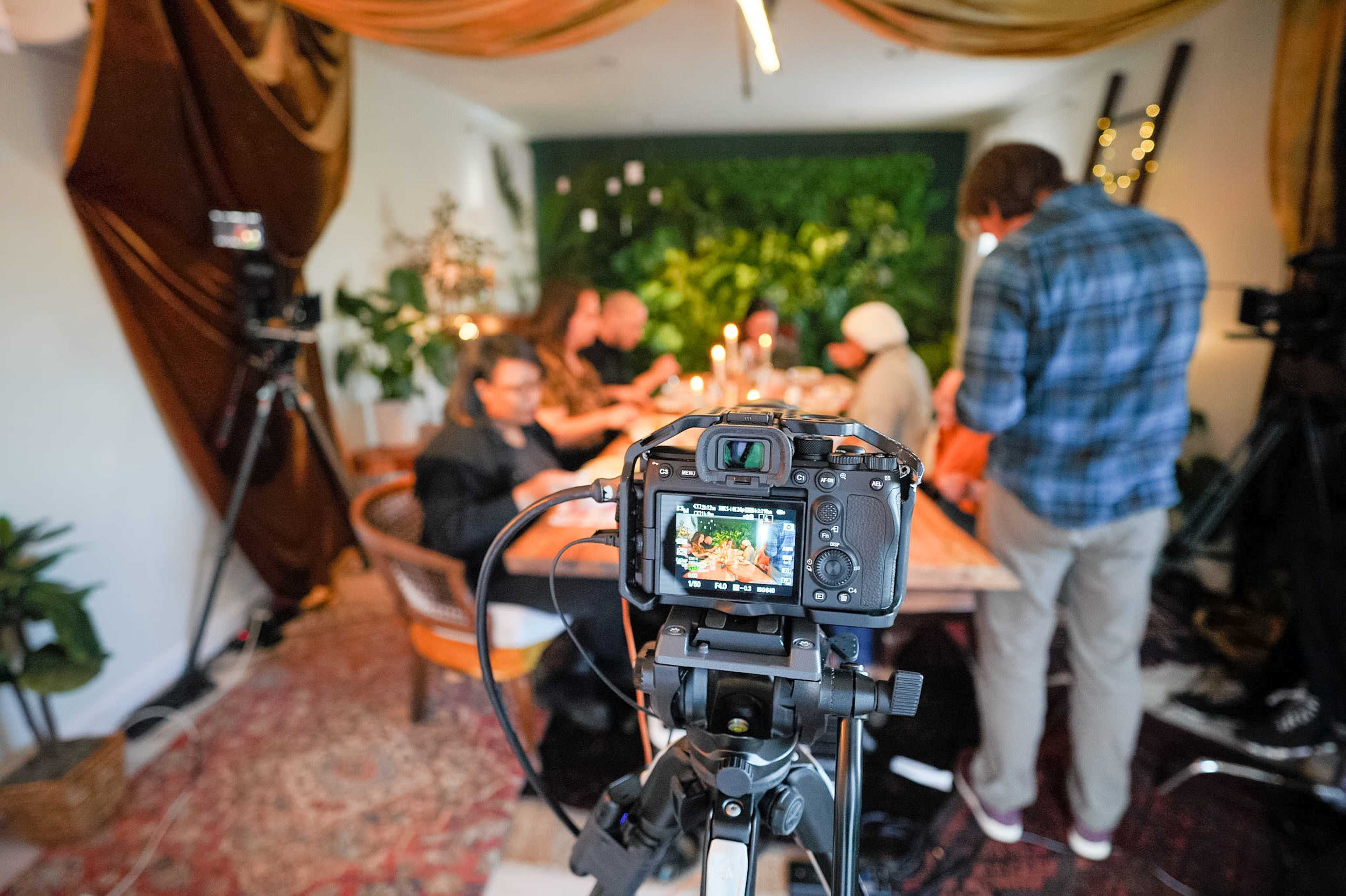 A camera on a tripod focuses on a dining table surrounded by people, with greenery and decorative drapes in the background.
