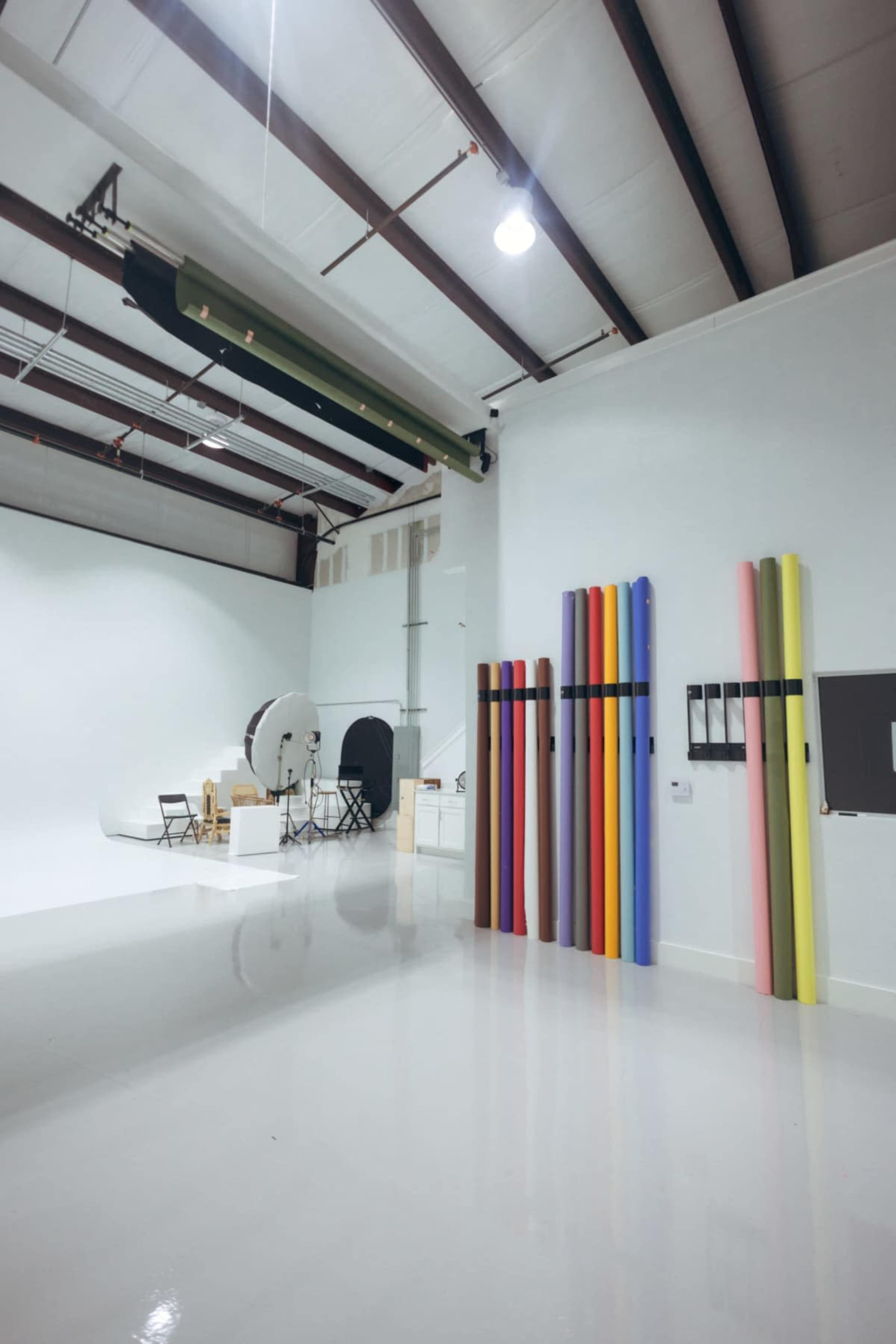 The image shows a photography studio with a white backdrop, various equipment, and colored paper rolls organized on a wall.