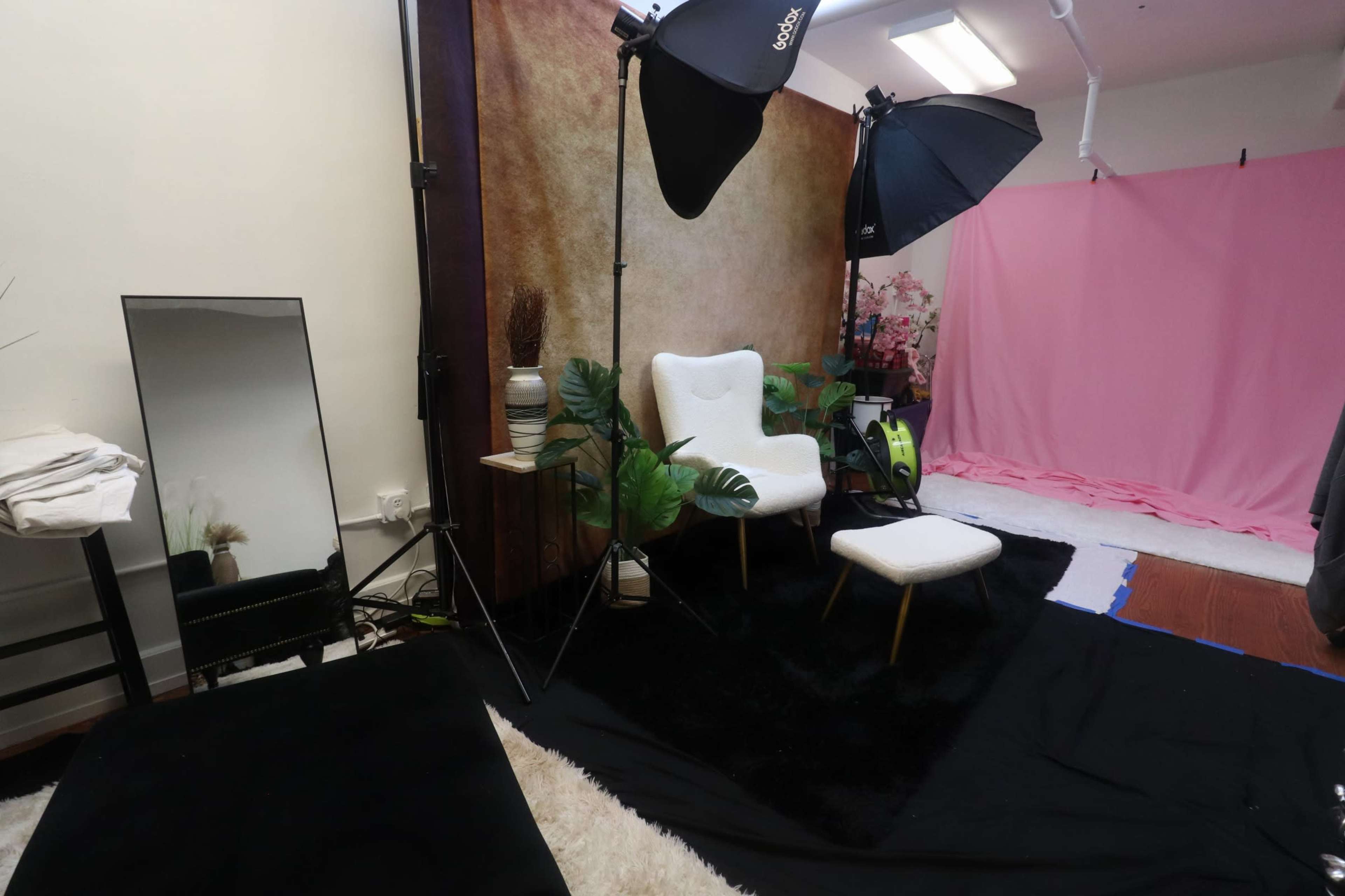 The image shows a photography studio setup featuring a black rug, white chairs, a small stool, and a pink backdrop with studio lights.