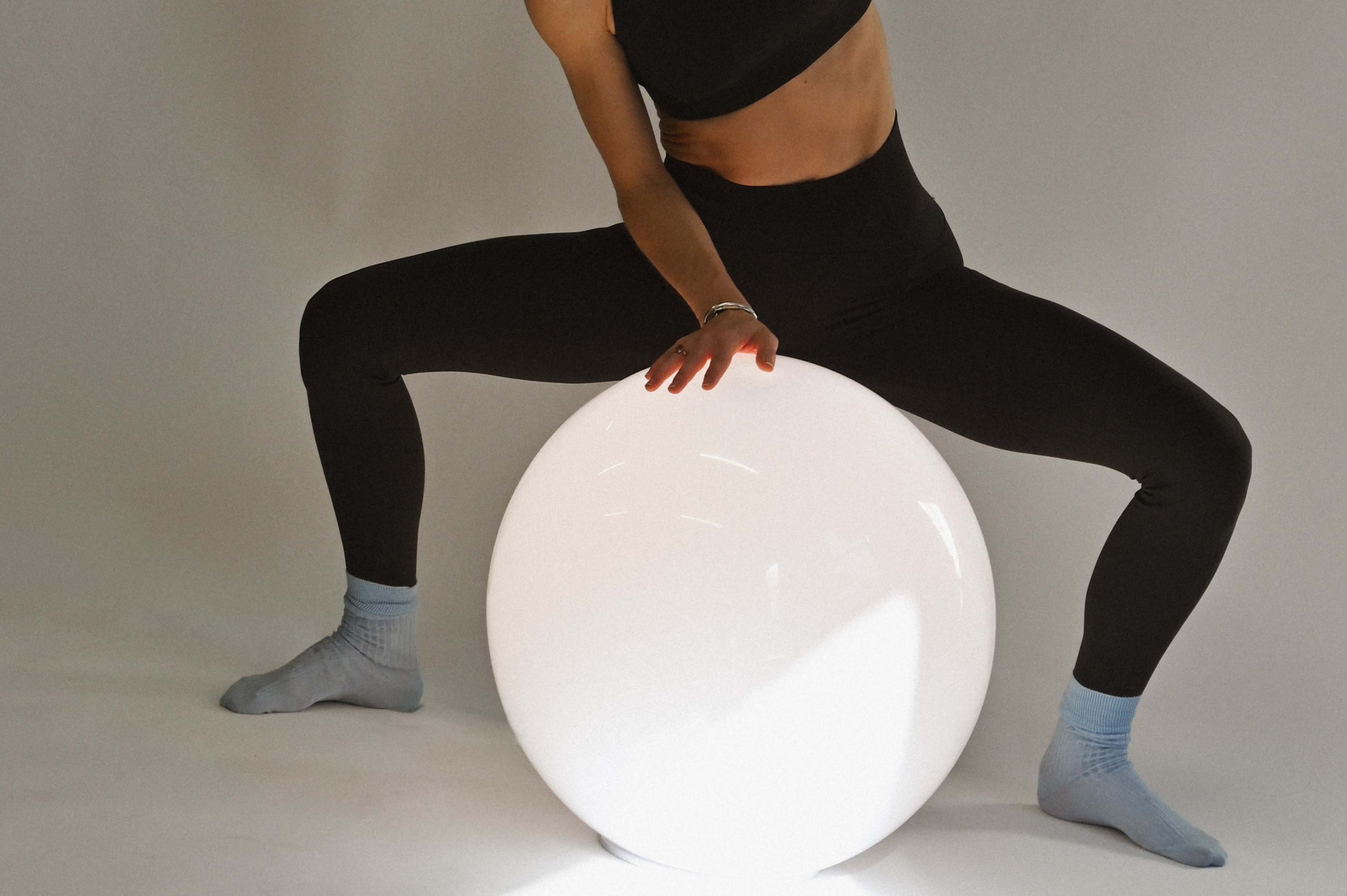A person in black attire and blue socks is crouching over a large, luminous white sphere against a plain background.