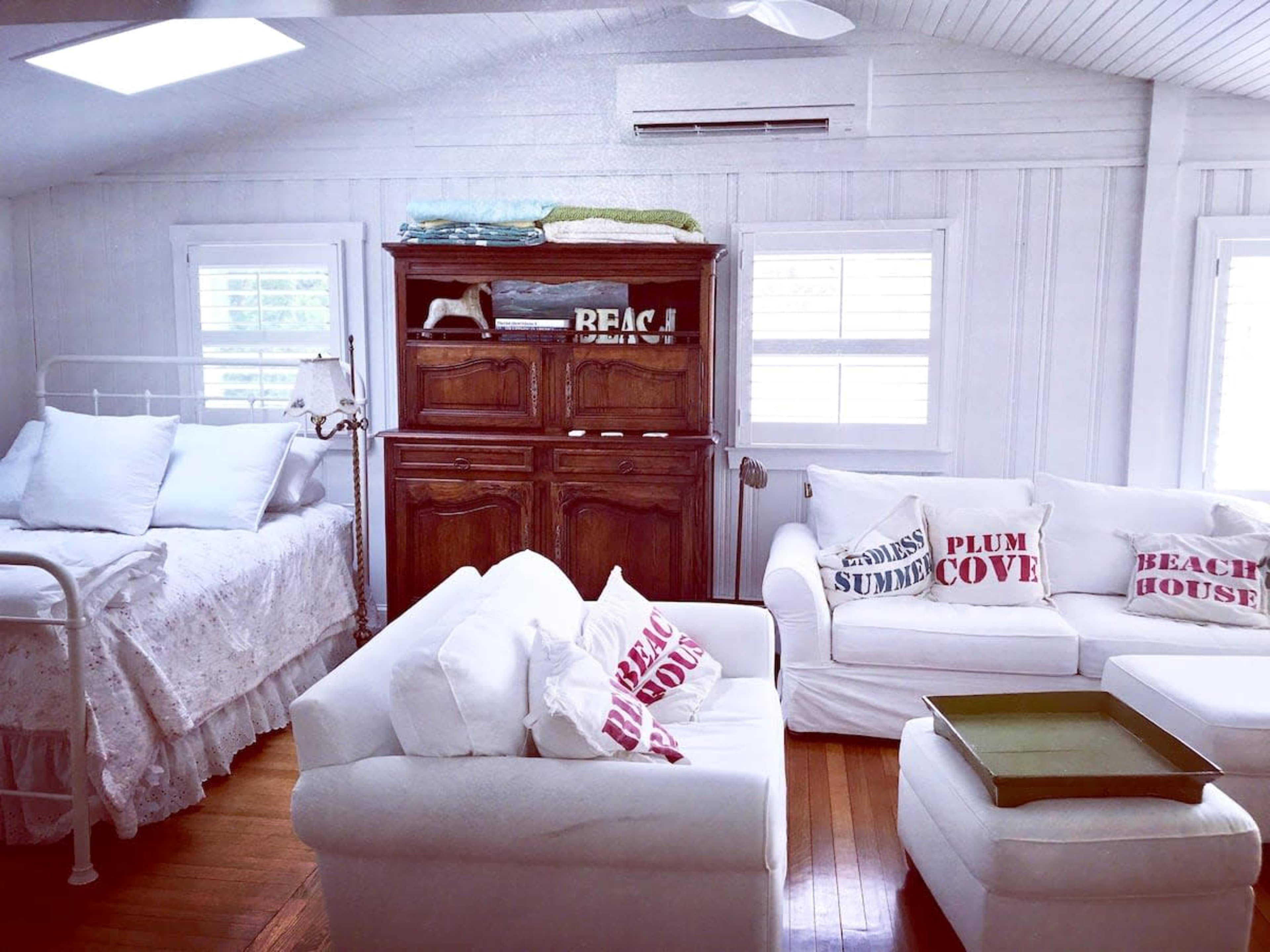 A bright, airy room with a white bed, a wooden storage unit, and two white sofas adorned with decorative cushions.