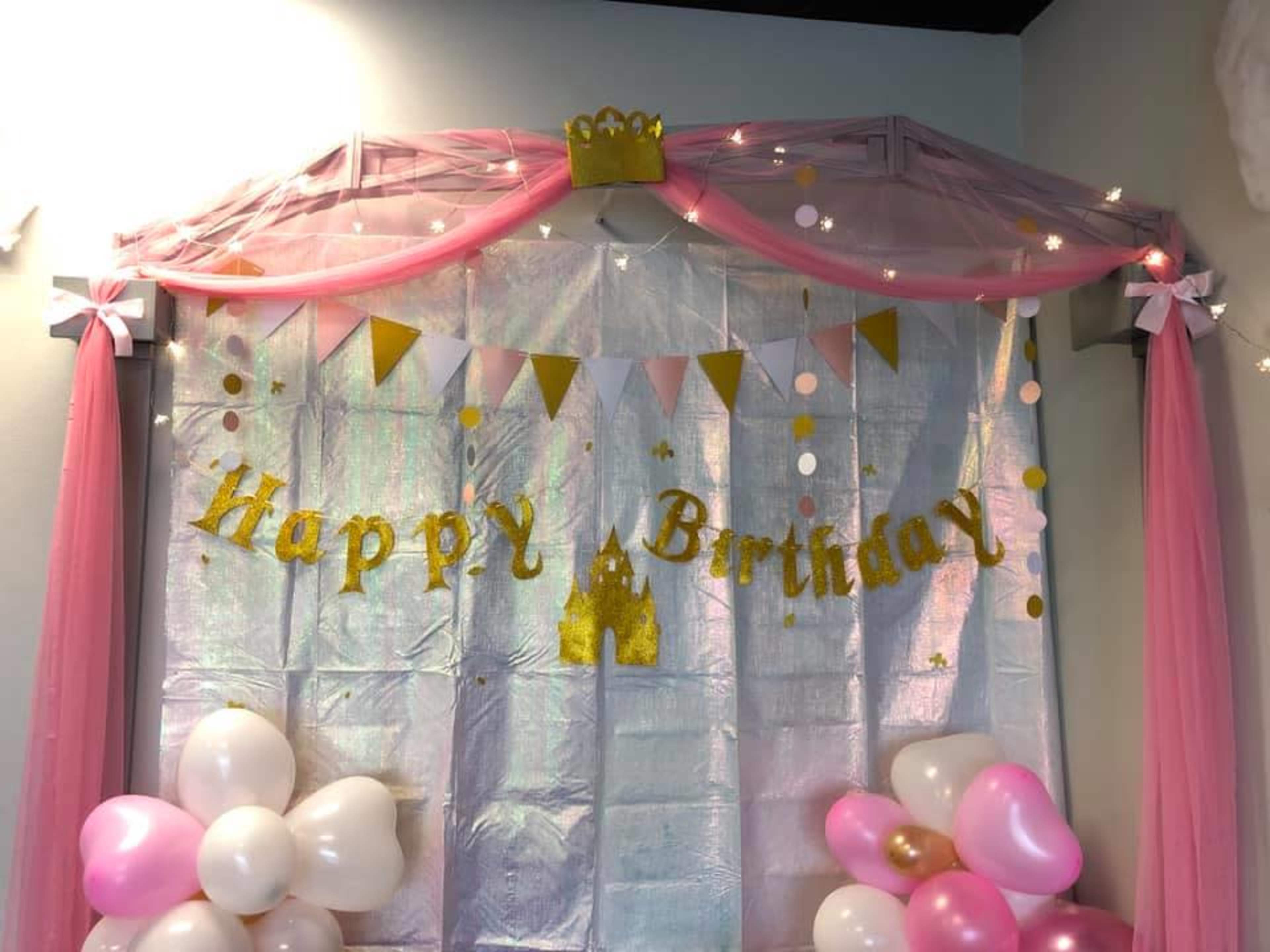 A colorful birthday party backdrop with a "Happy Birthday" banner, pink and gold decorations, and balloons.