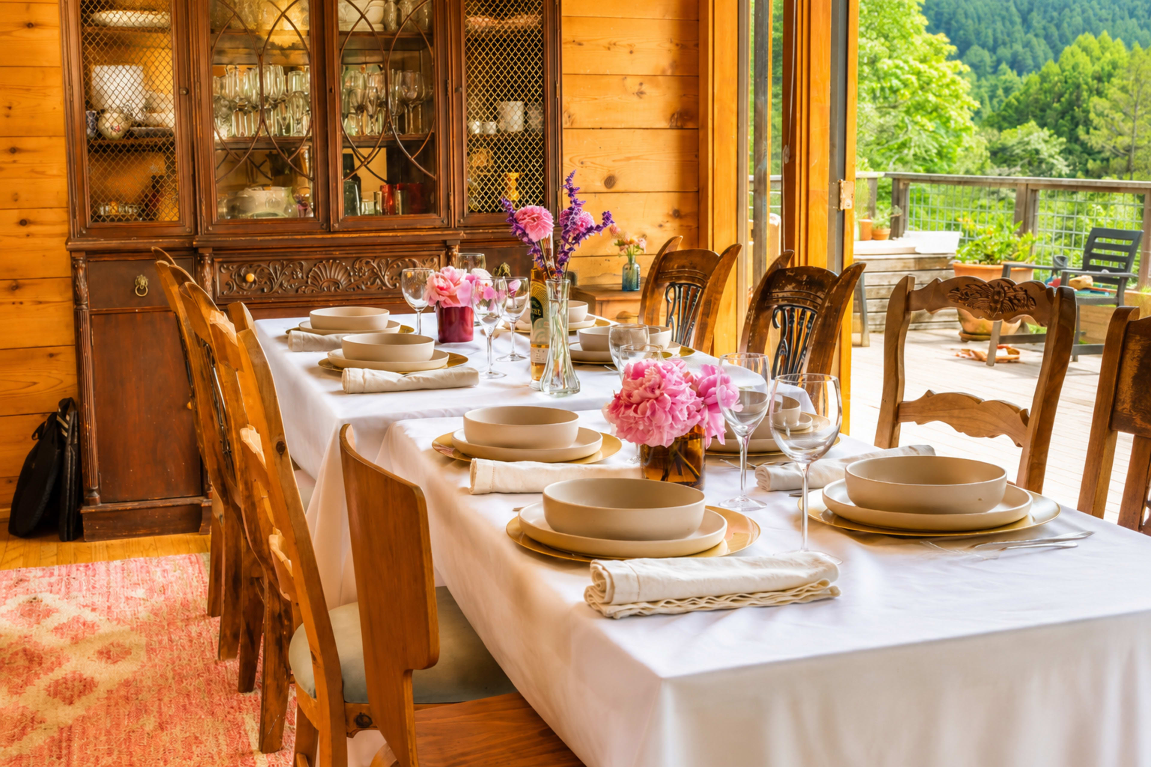 A long dining table is set with plates, glasses, and floral centerpieces, surrounded by wooden chairs in a well-lit room with large windows overlooking greenery.