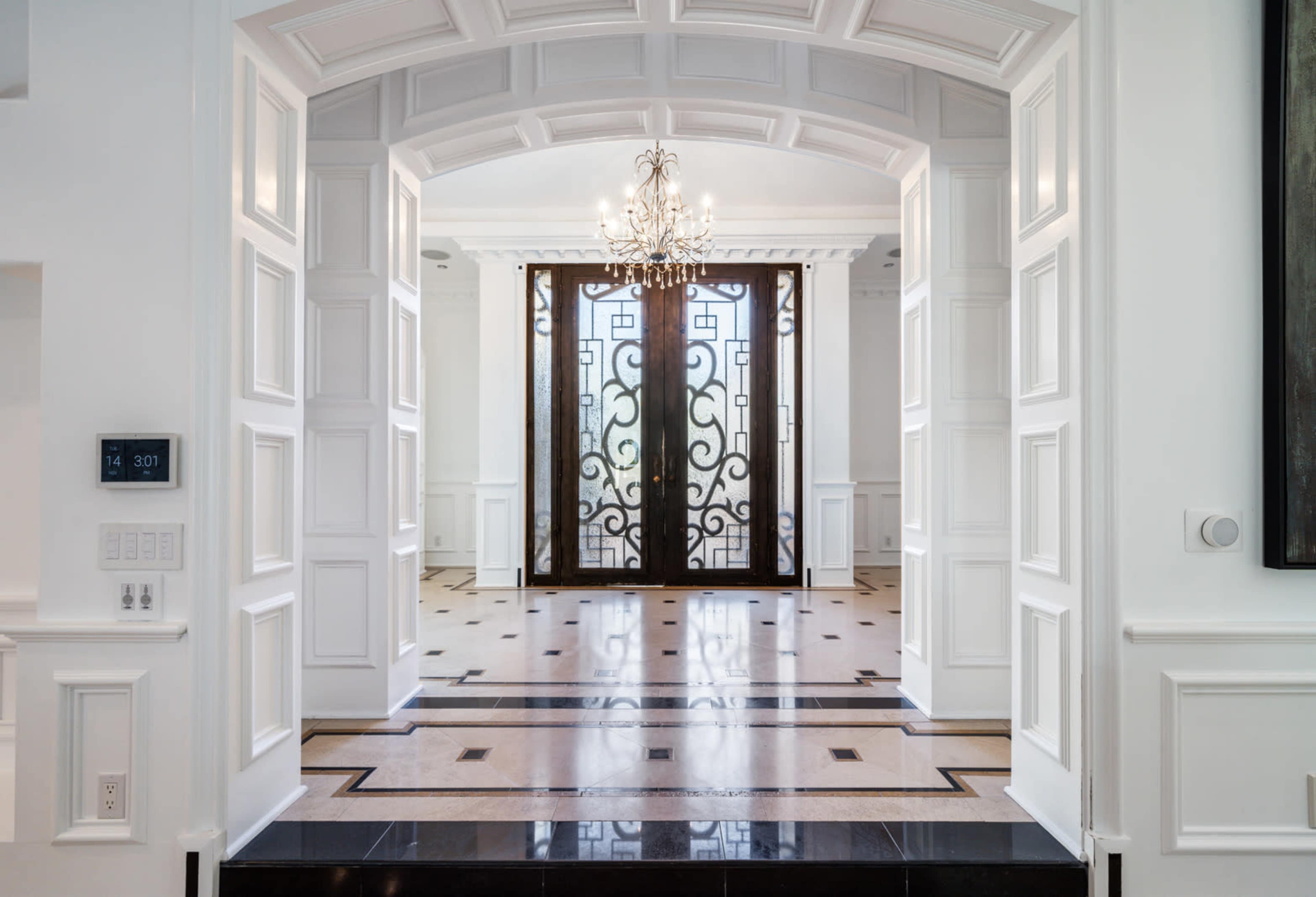 An elegant entryway featuring a large decorative doorway with a chandelier, framed by white paneling and a marble floor.