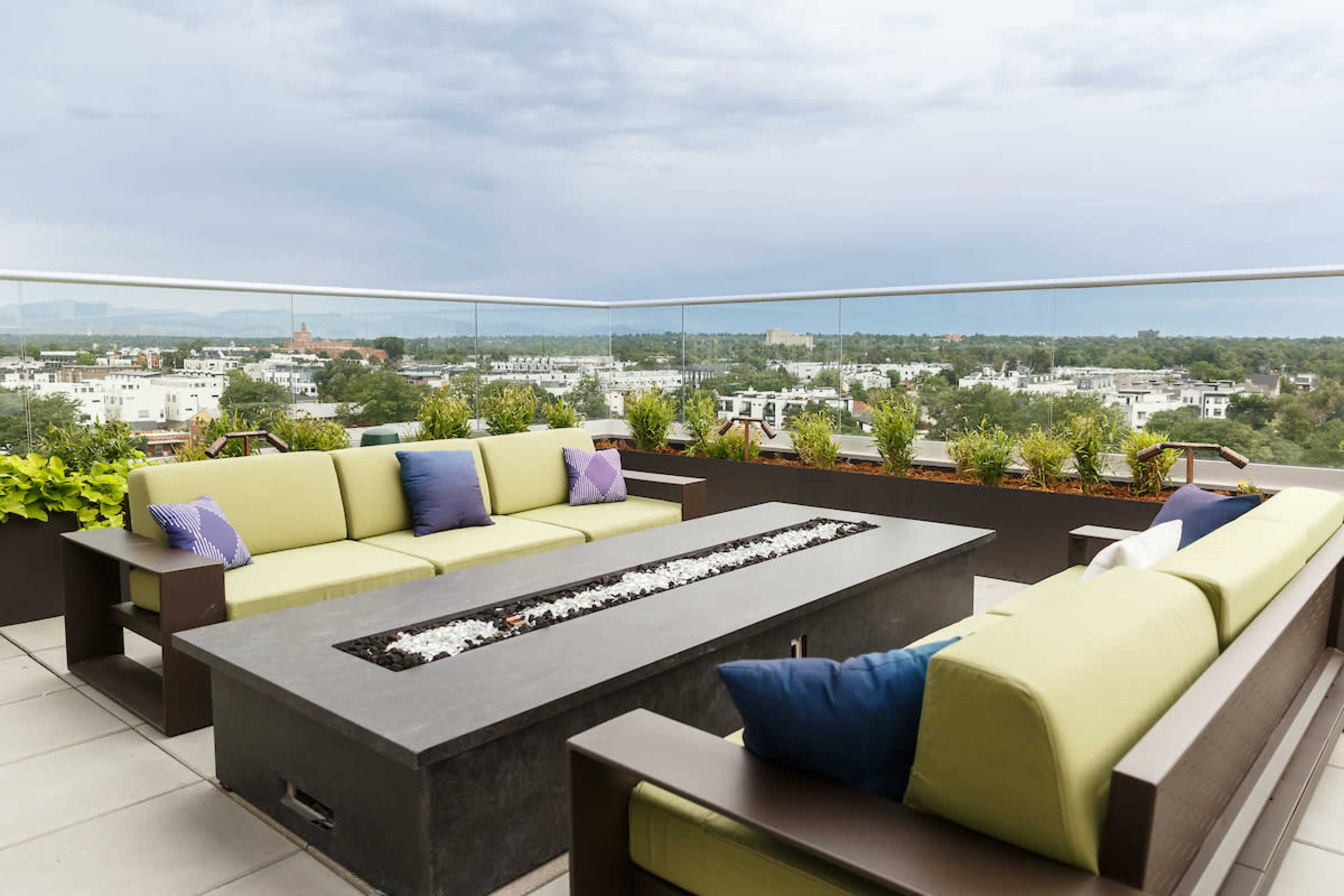 A rooftop lounge area features green sofas with decorative pillows arranged around a rectangular fire pit, overlooking a cityscape.
