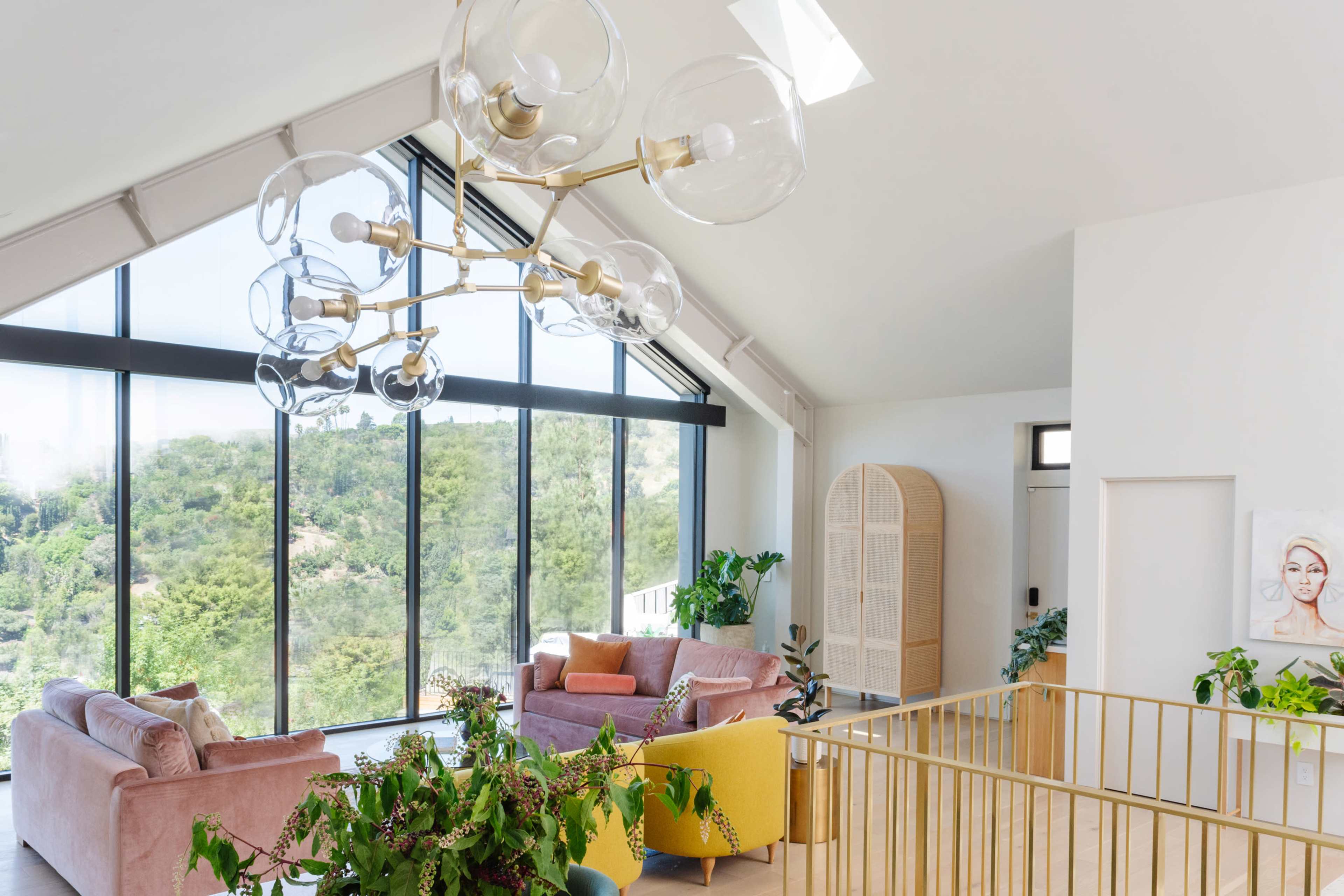 A modern living room features large windows with a view of greenery, a chandelier with glass orbs, and a mix of pink and yellow furniture.