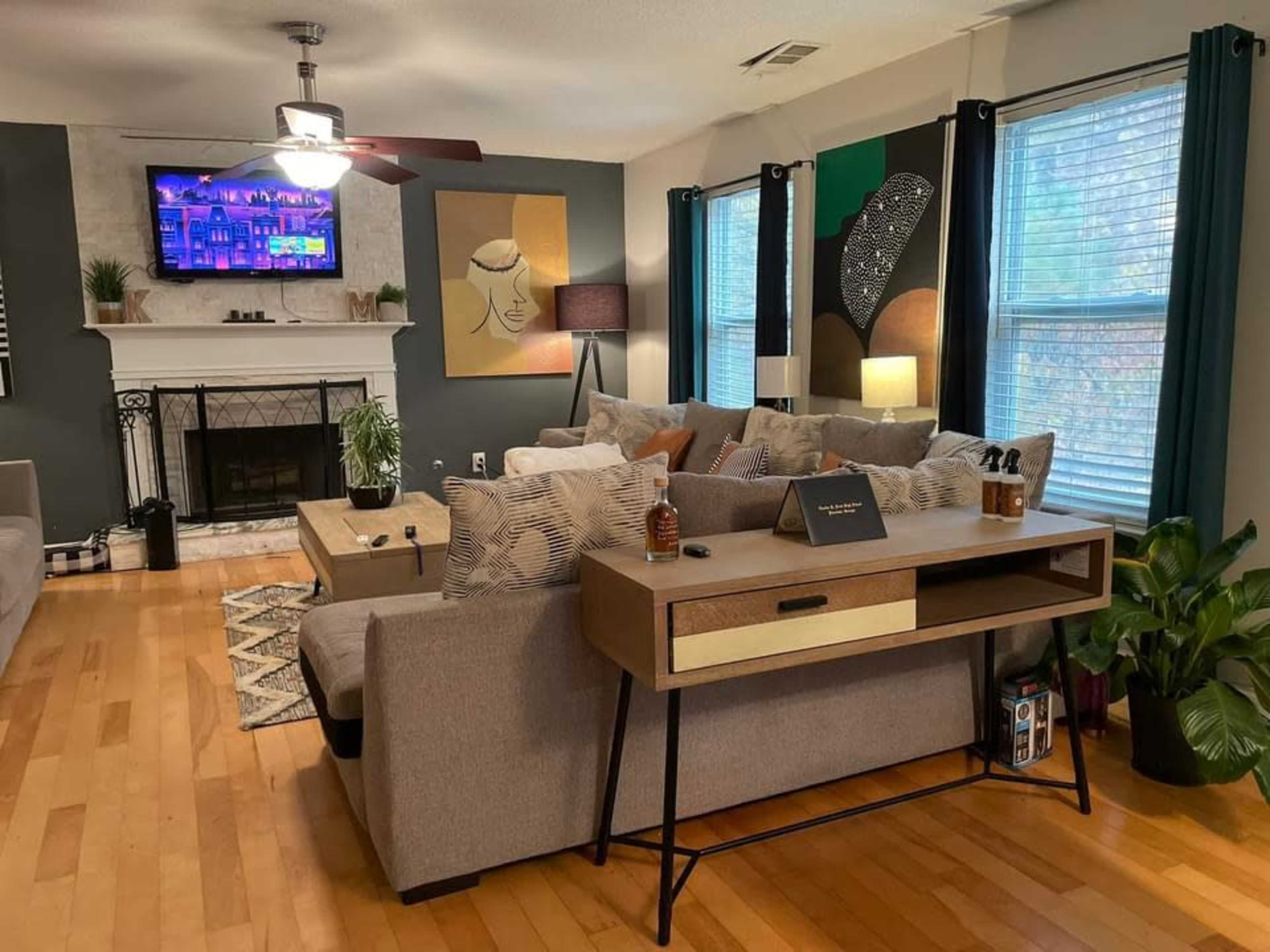 A living room with a comfortable sofa arranged around a coffee table, a fireplace, a mounted TV, and a modern desk under a window.