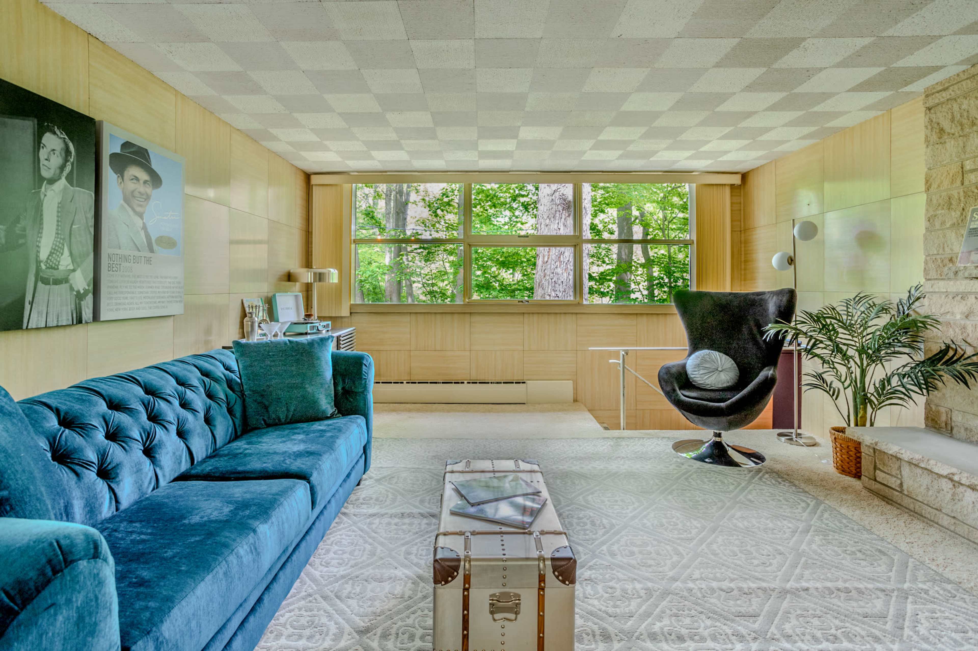 A modern interior living space featuring a blue tufted sofa, a black chair, a glass coffee table, and large windows with views of greenery outside.