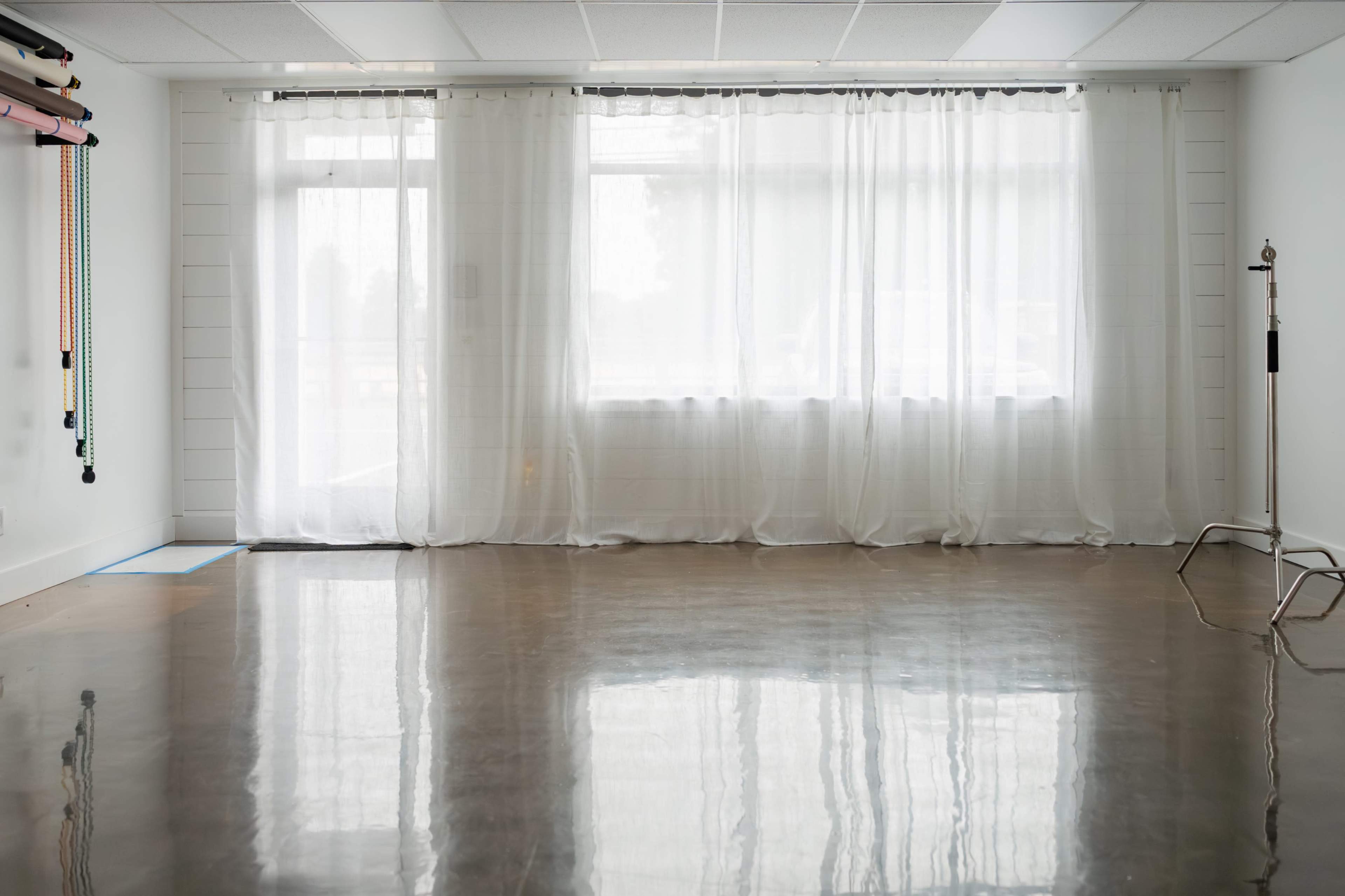 The image shows a minimalist interior space with a polished concrete floor, large windows covered by sheer white curtains, and studio equipment along one wall.