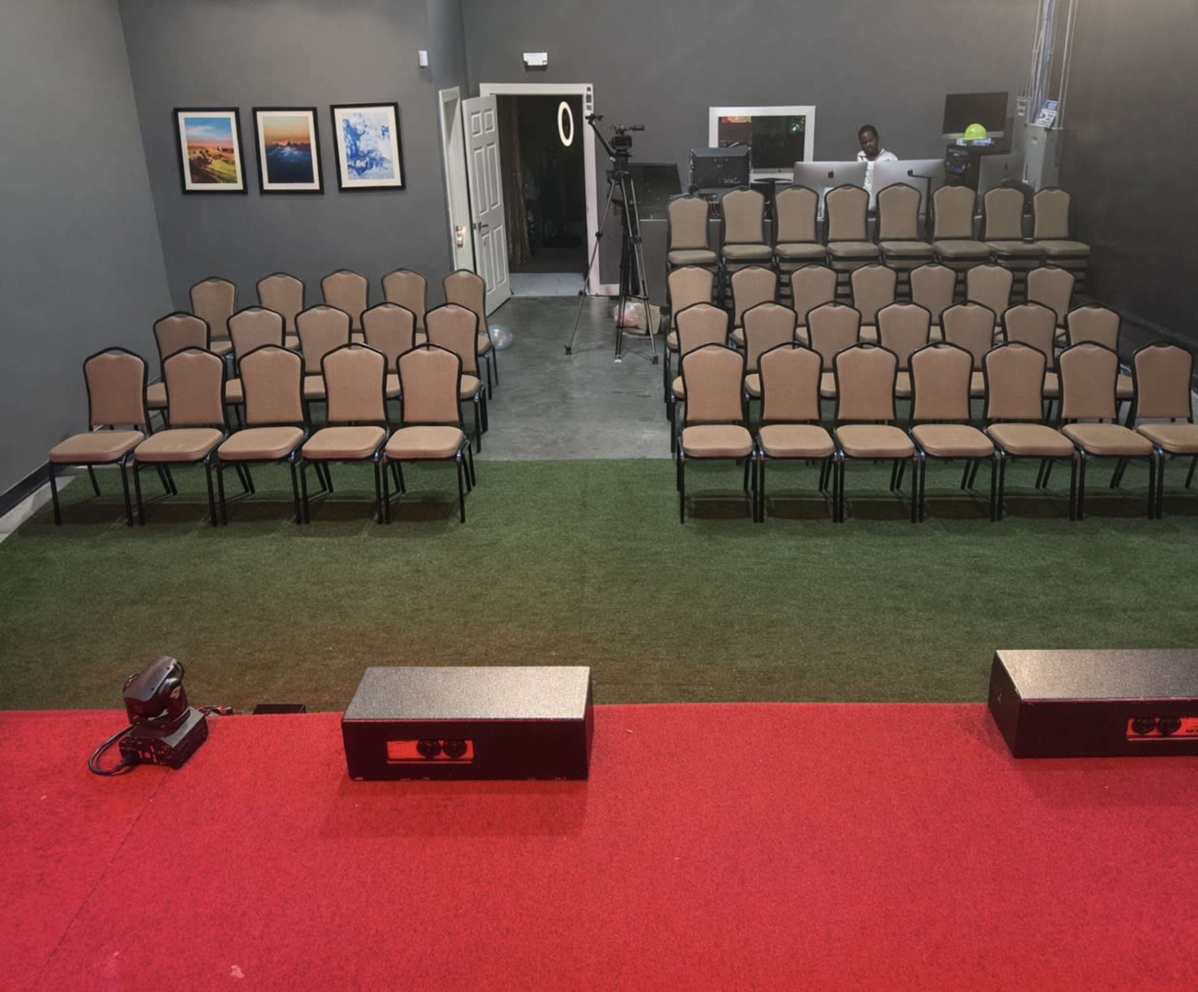 The image shows a seating area with rows of empty chairs facing a stage, set against a gray wall with framed pictures, and a small control area visible in the background.