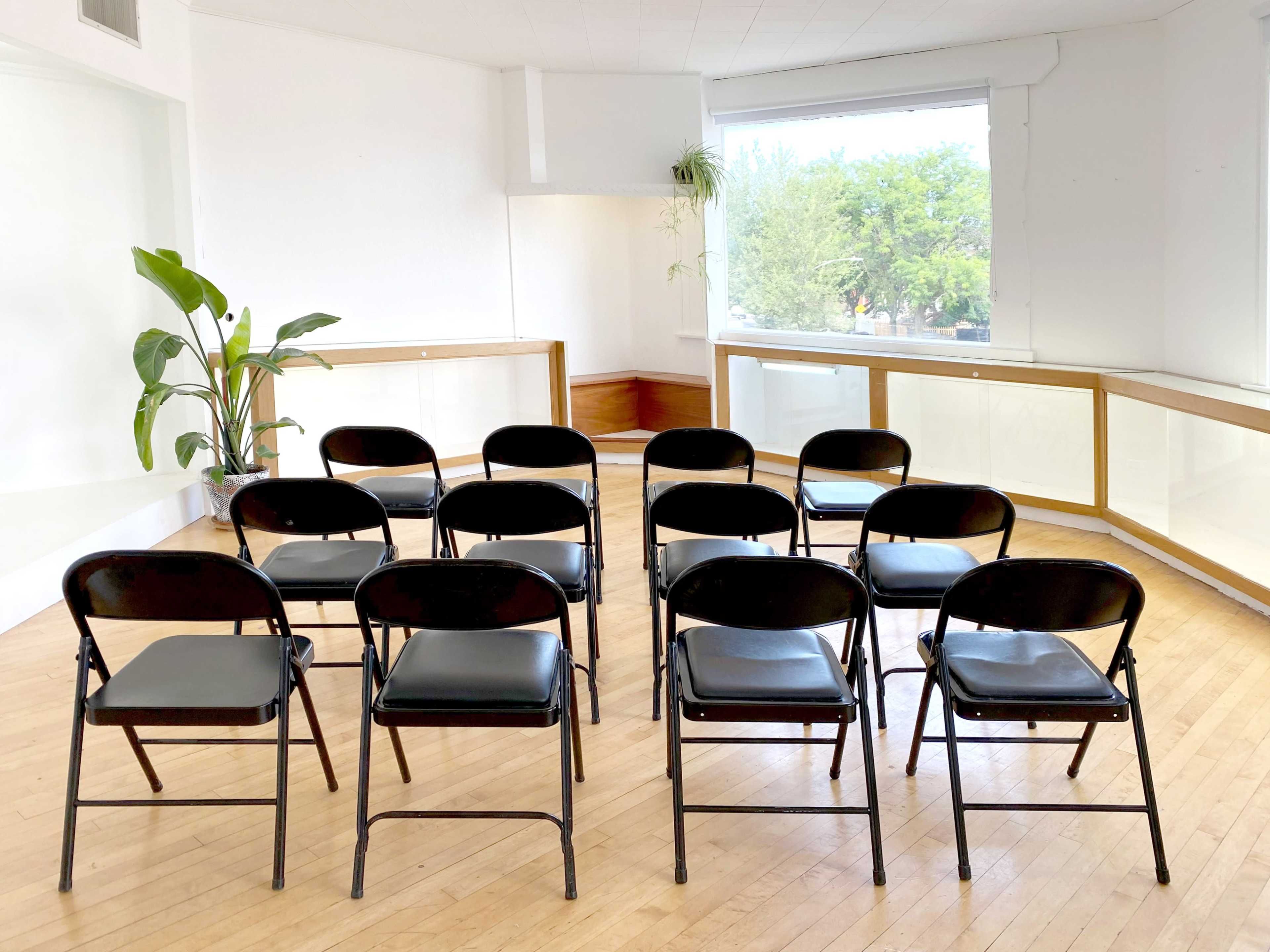 A group of black folding chairs is arranged in two rows facing a large window in a bright, empty room.