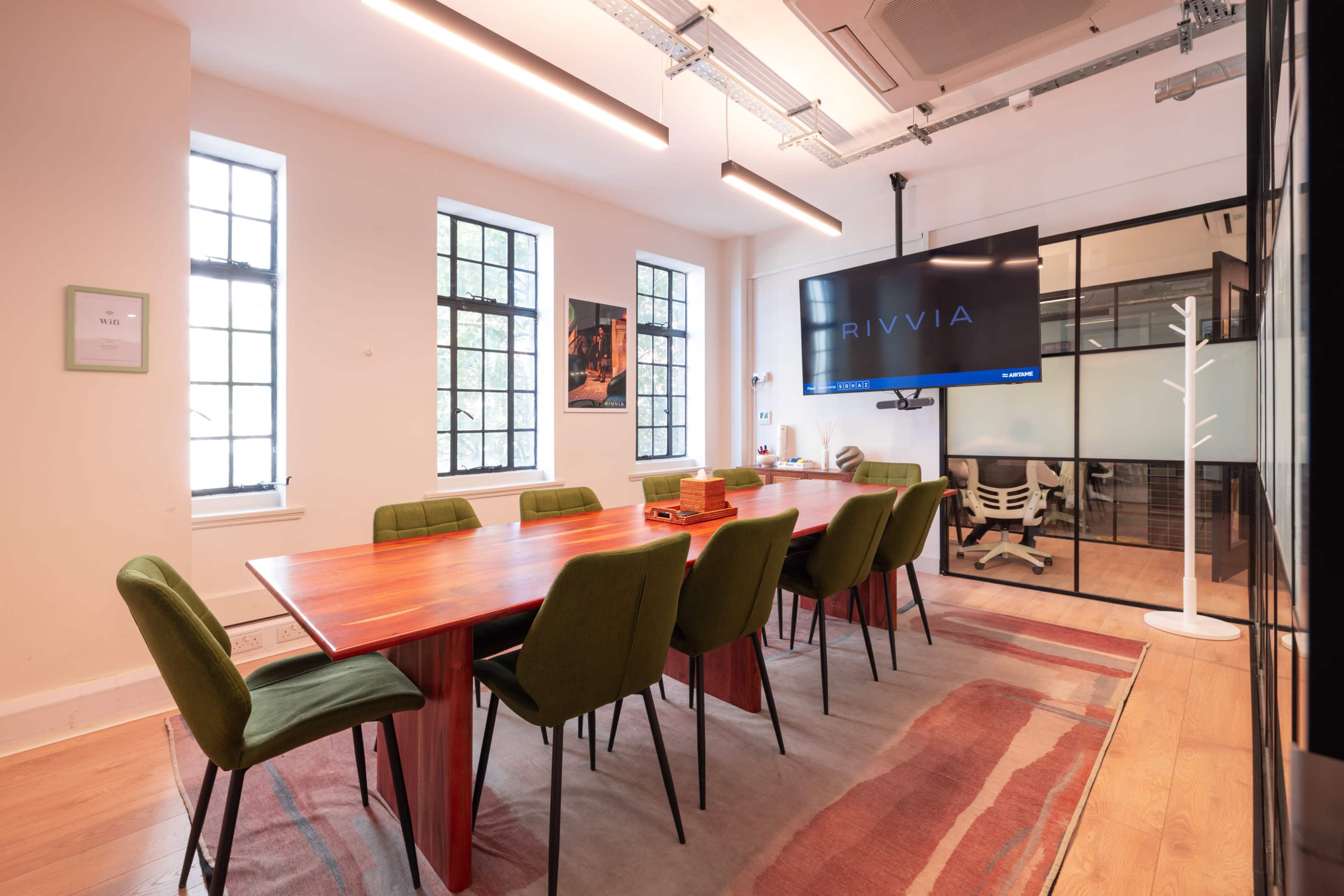 A modern meeting room features a long wooden table surrounded by green chairs, a large screen, and multiple windows for natural light.