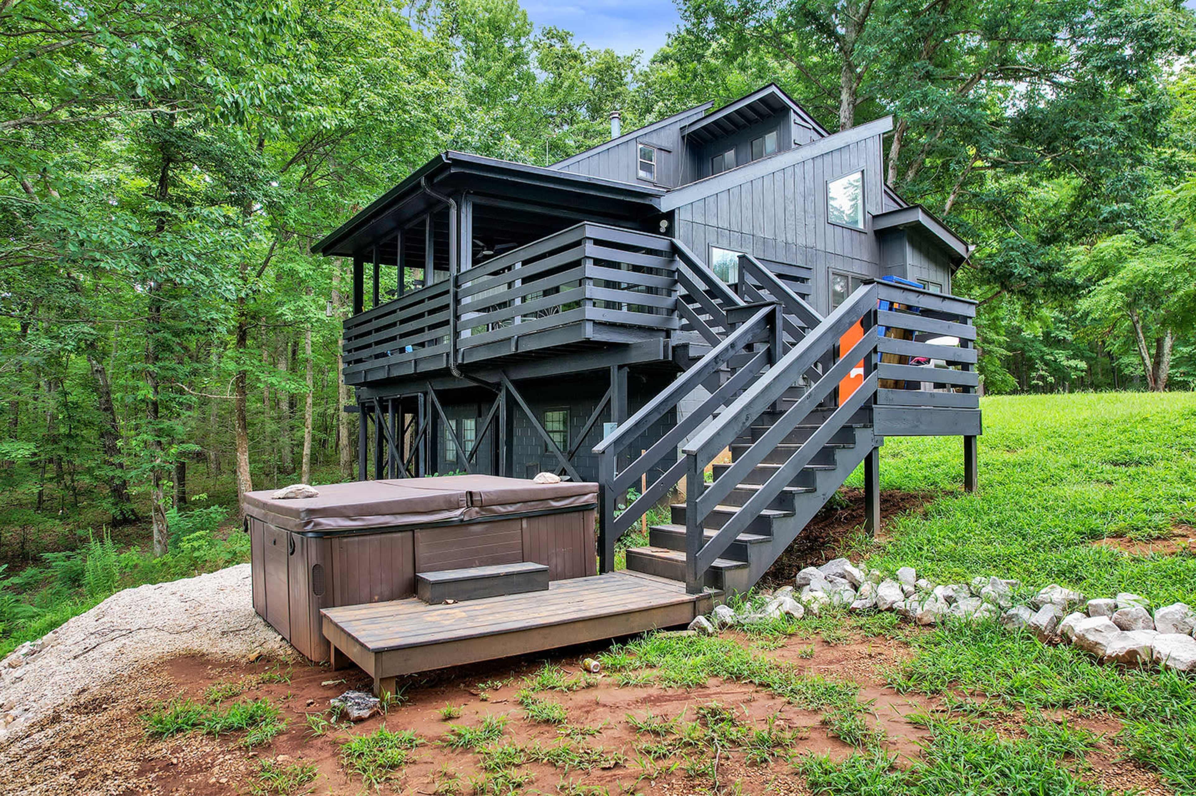 A two-story black house with a deck and hot tub is surrounded by greenery and trees.
